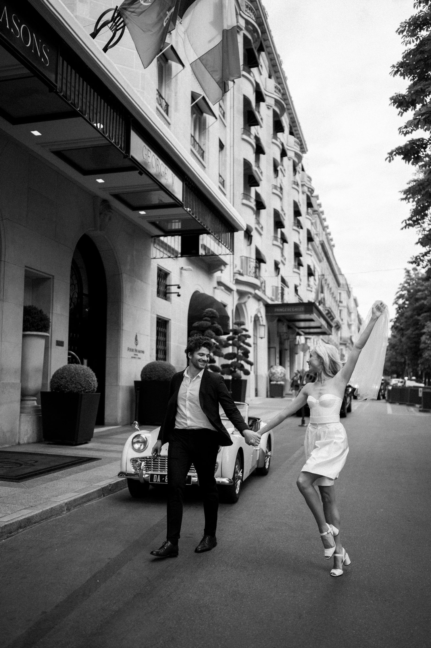 Black and white photo of newlyweds dancing outside luxury Paris hotel with vintage convertible car.