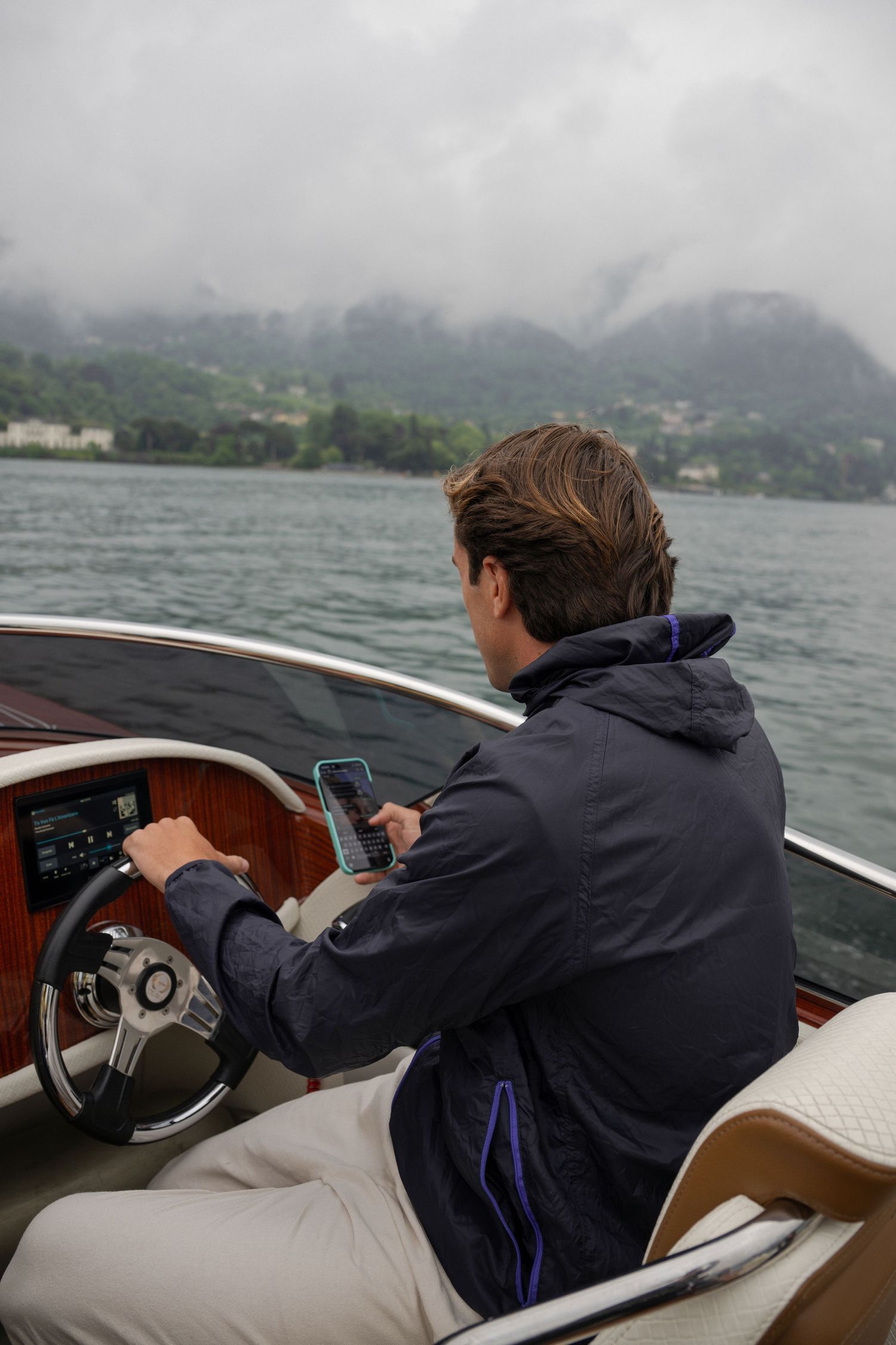 Boat captain navigating a classic wooden speedboat on a cloudy mountain lake while checking his phone.