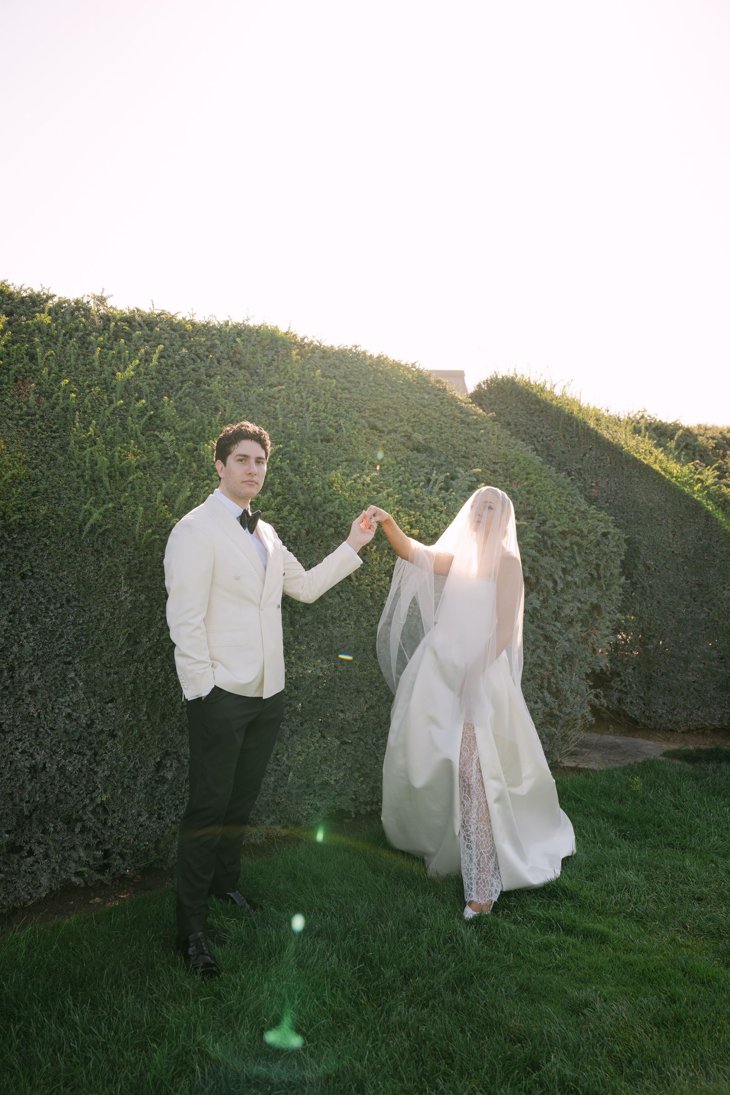 Wedding couple holding hands near tall green hedges, groom in ivory jacket, bride in lace wedding dress with veil.