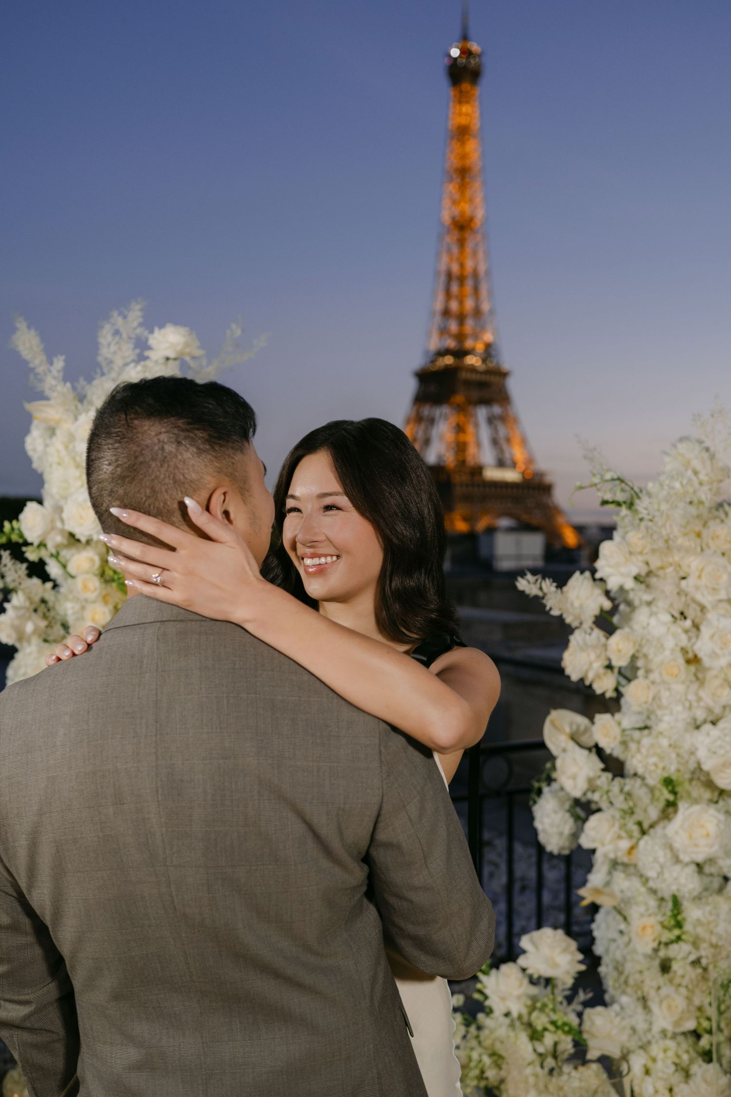 Smiling woman wraps her arms around a man in a suit during a Paris proposal with white flowers and the Eiffel Tower at dusk.