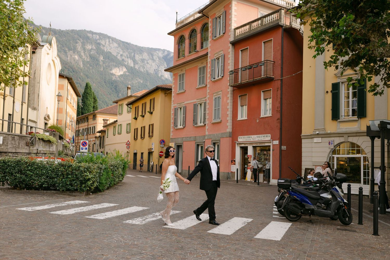 Couple in formal attire crossing a zebra crossing in a charming Italian town square with colorful buildings and mountains.
