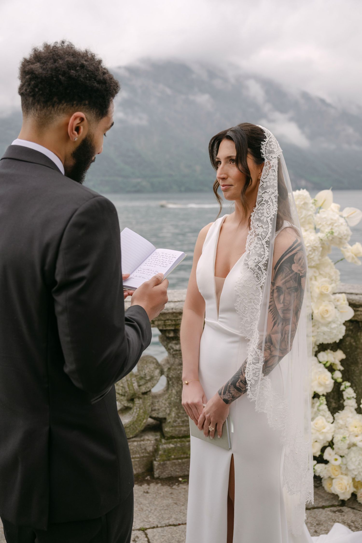 Groom reads vows to tattooed bride in white gown and lace veil at lakeside wedding ceremony.