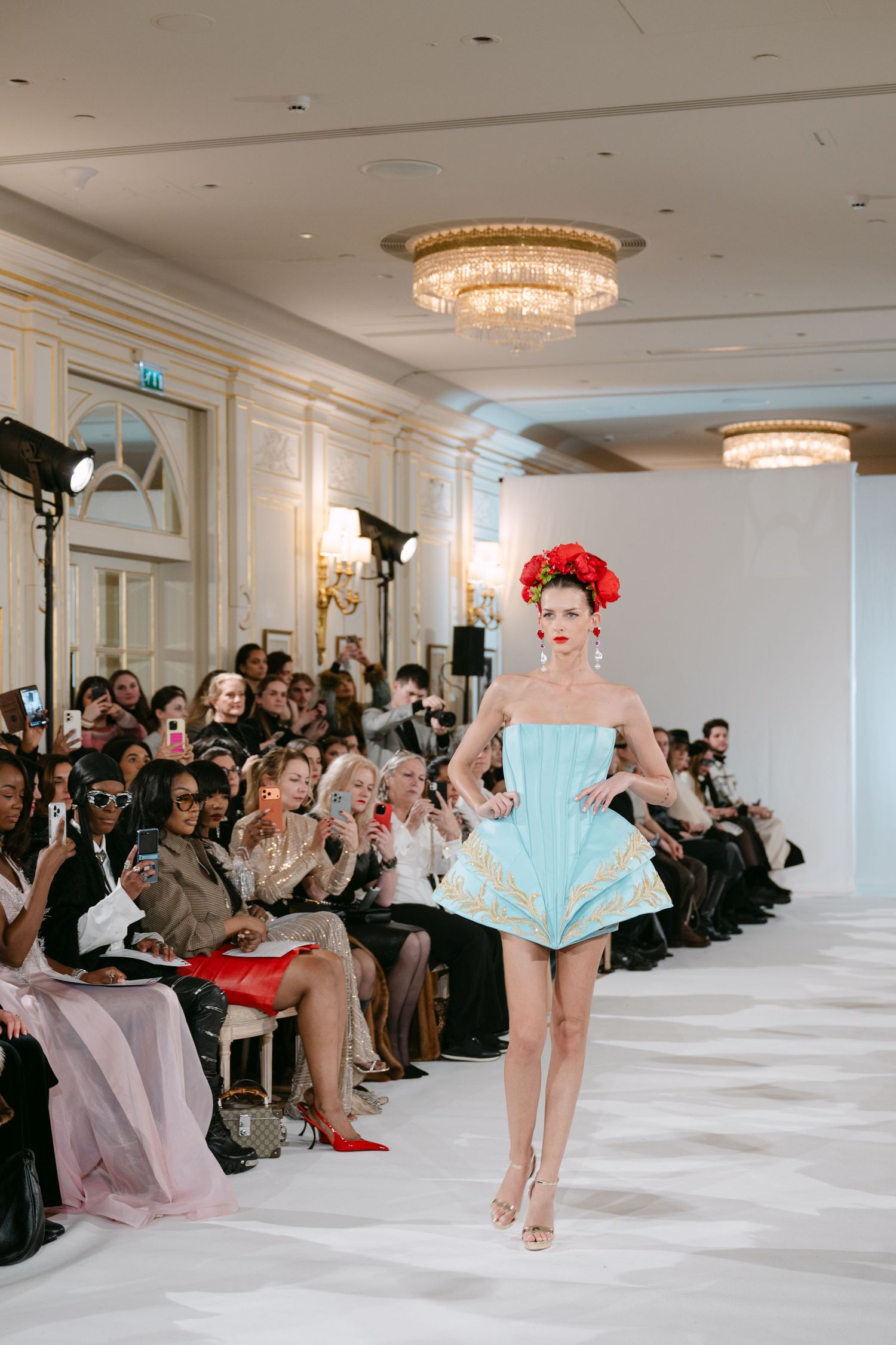 Model in light blue strapless mini dress with red floral crown walks couture runway.
