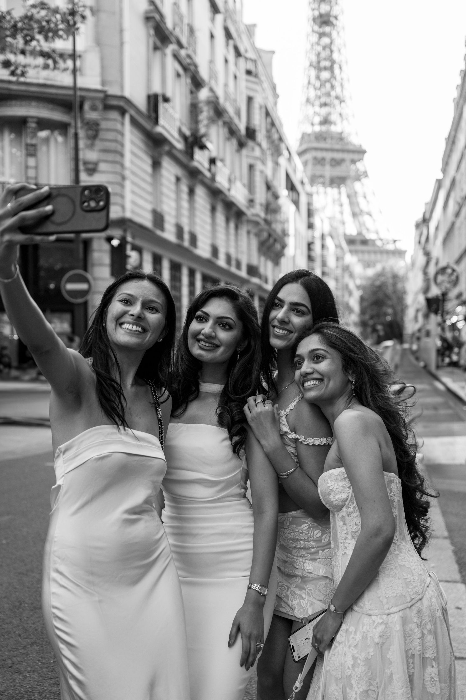 Four women in white dresses take a selfie on a Paris street with the Eiffel Tower behind them.
