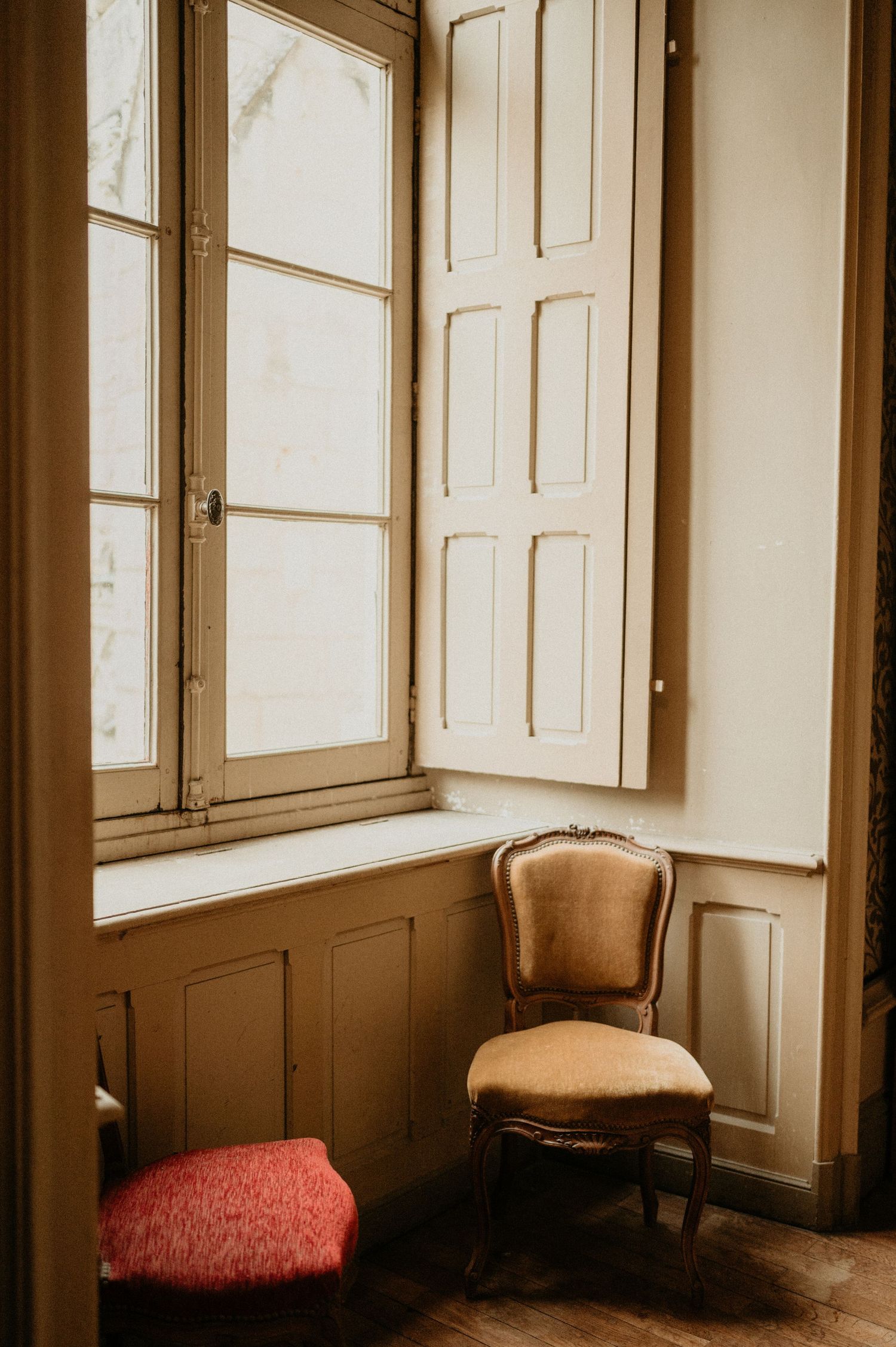 Minimalist corner with vintage chair by window featuring ivory shutters and red cushion.