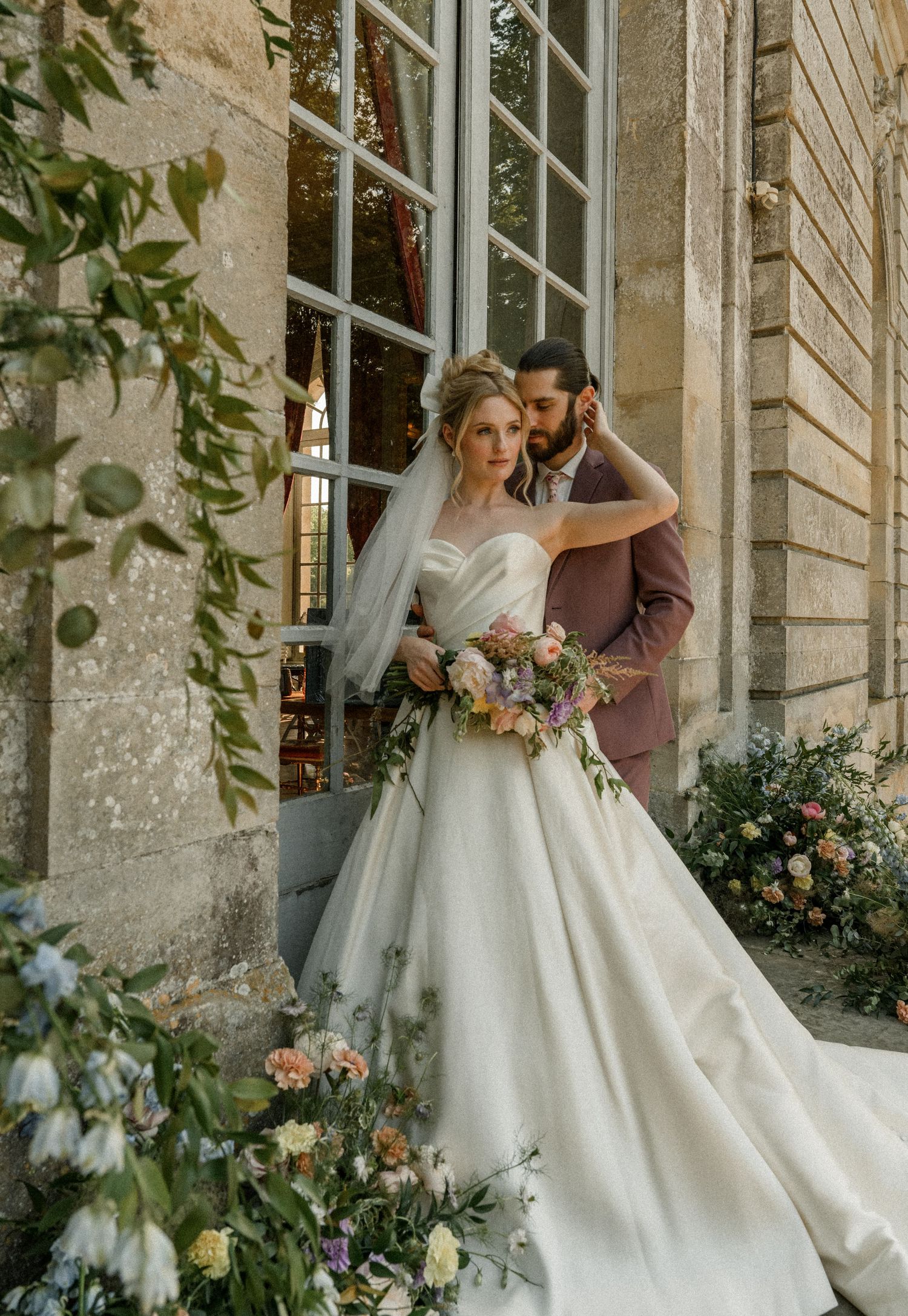 A wedding couple shares an intimate moment against an old stone building surrounded by climbing flowers.