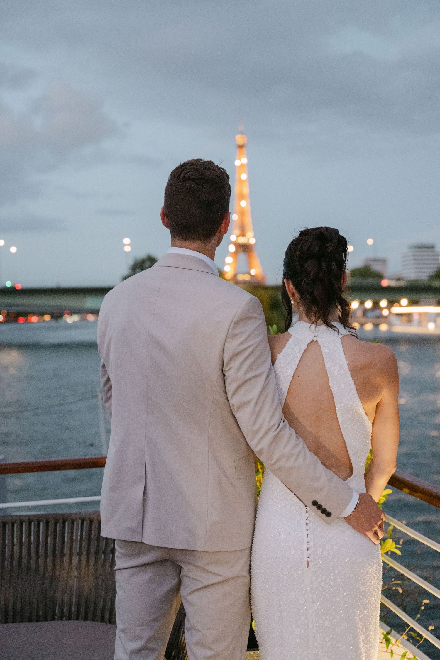A couple in wedding attire stands at the railing of a boat deck overlooking city lights at dusk.
