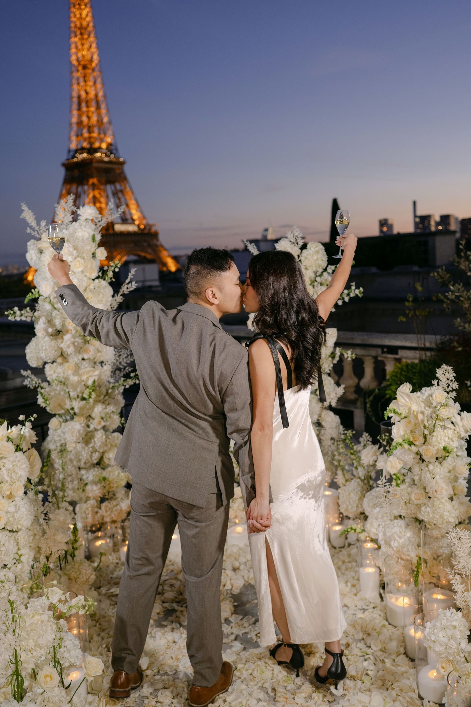 Couple kisses on a Paris rooftop surrounded by white roses and candles, with the illuminated Eiffel Tower glowing at sunset.