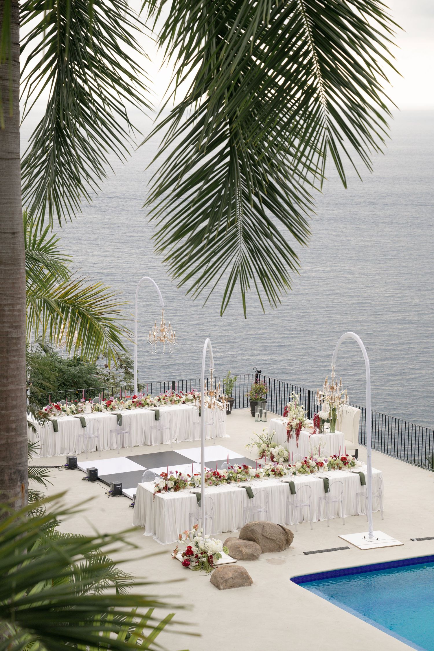 Elegant outdoor wedding reception setup with white tables, floral arrangements, and ocean views framed by tropical palm trees.