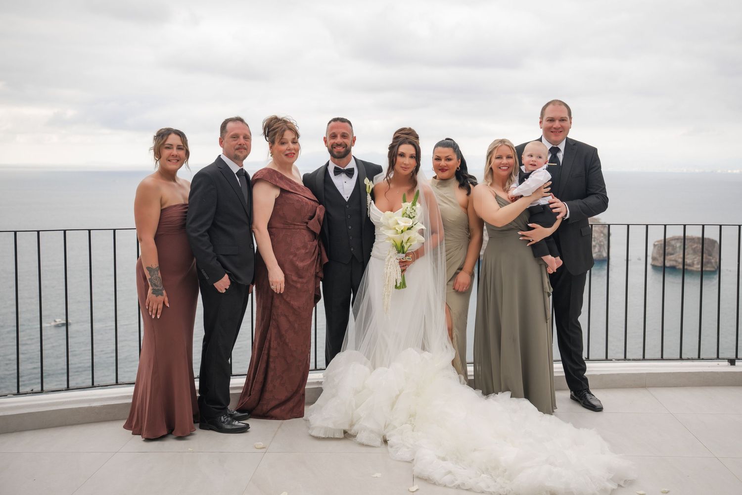 Wedding party poses on scenic coastal terrace, bride in white gown with bouquet, groom in suit, family in formal attire.
