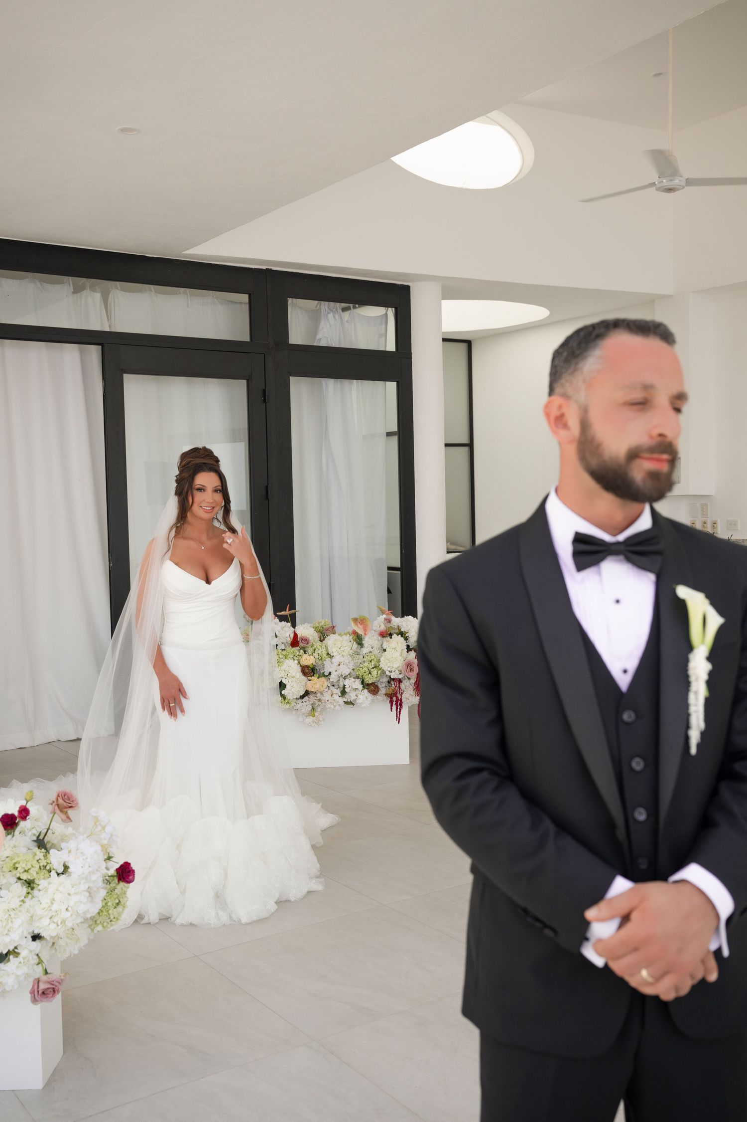 A groom in a black tuxedo stands in the foreground as his bride in a white gown and veil approaches for their first look.