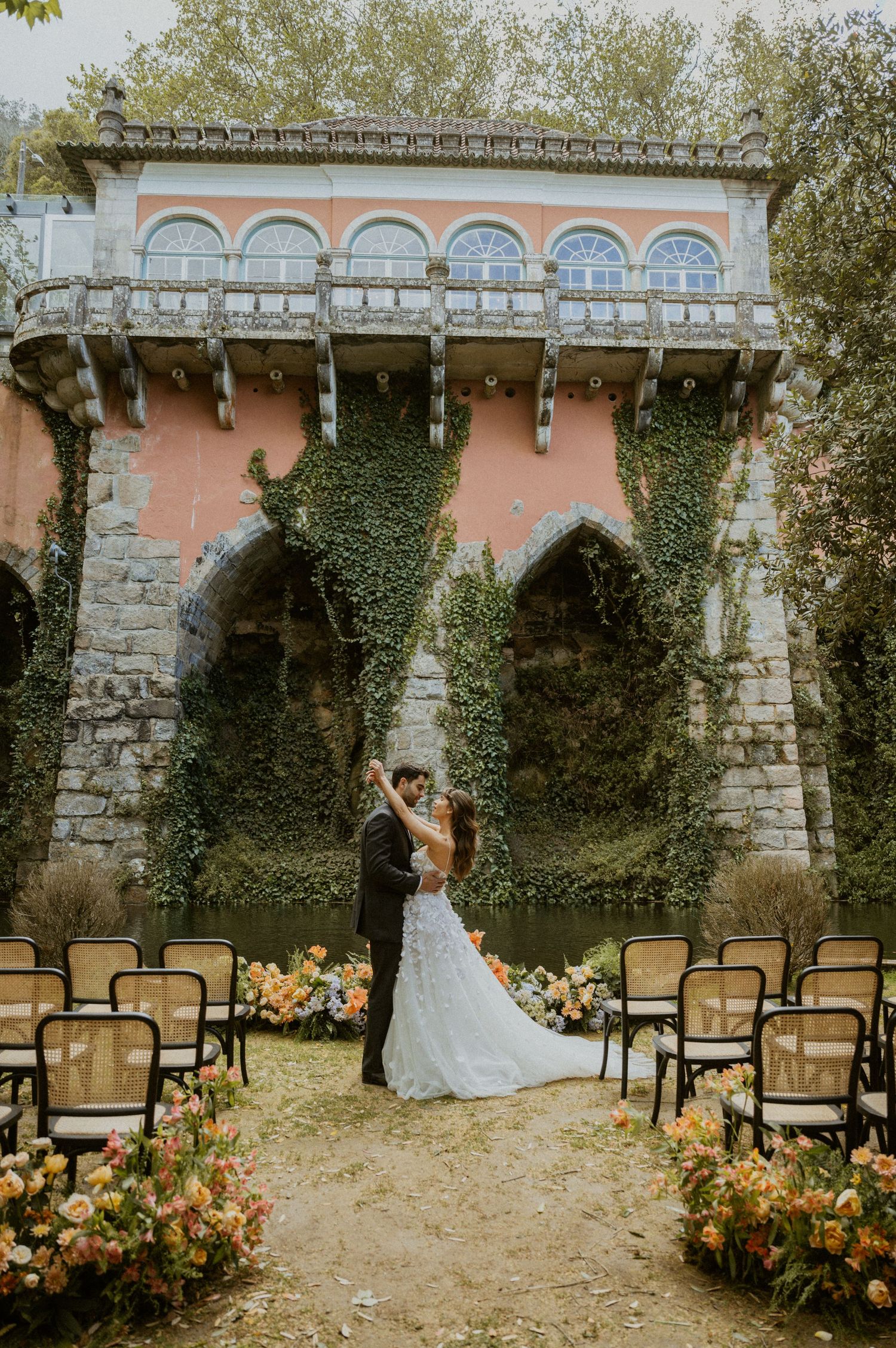 Bride and groom embrace at outdoor wedding ceremony with ivy-covered pink villa and floral arch aisle.