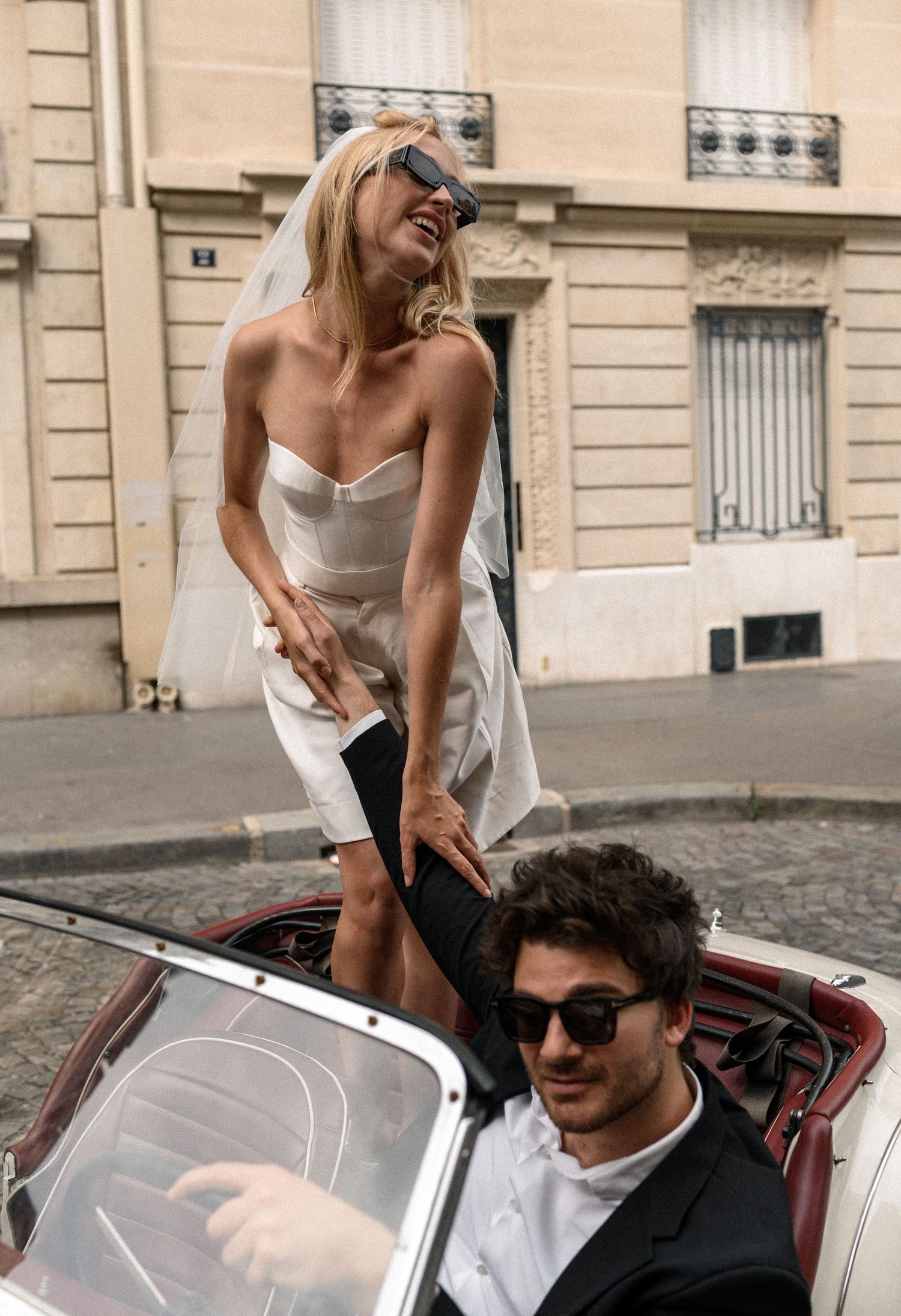Bride in white mini dress and veil stands in vintage convertible while groom in black suit holds her hand in Paris.