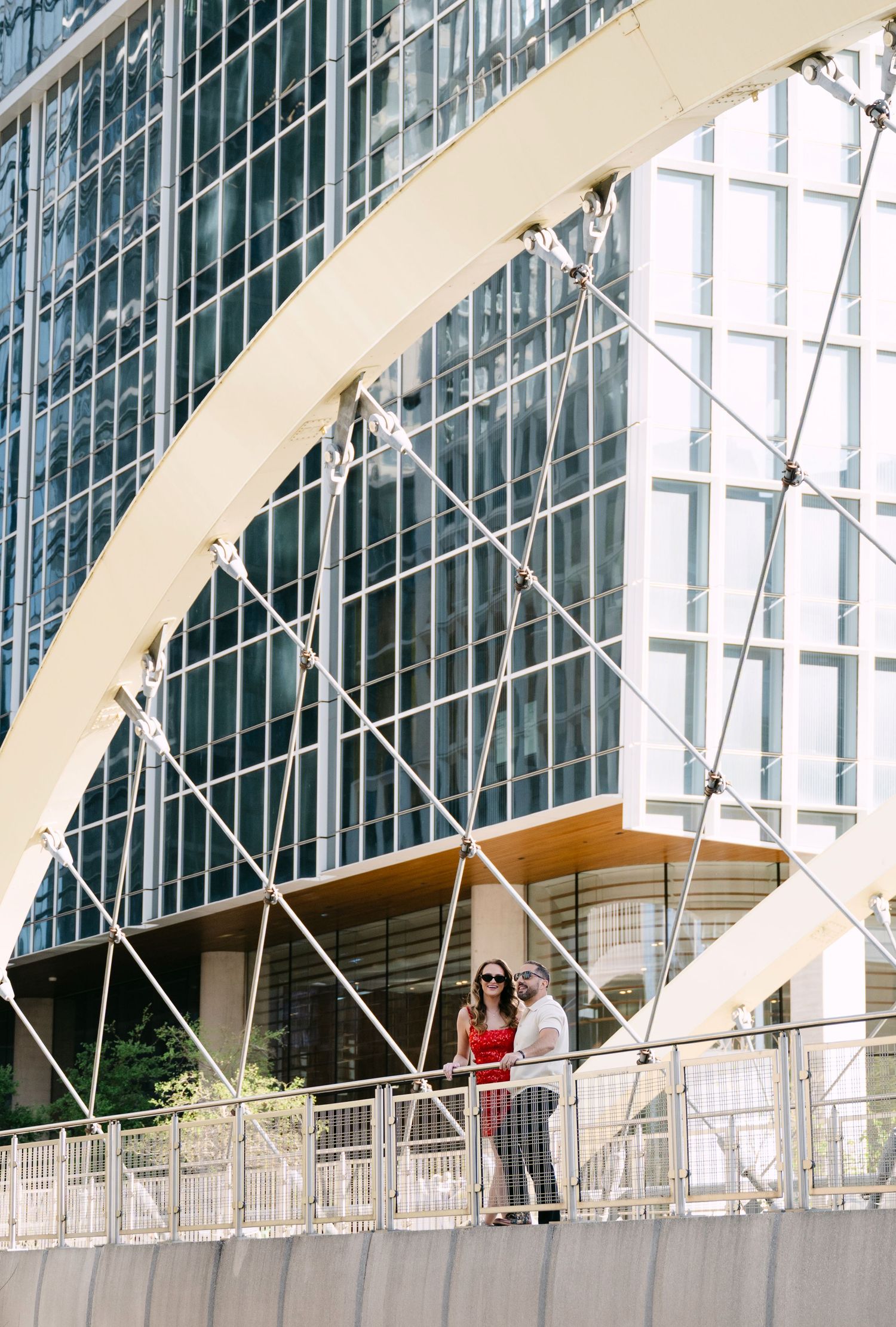 Couple poses together on pedestrian bridge with modern glass skyscraper architectural backdrop behind them.