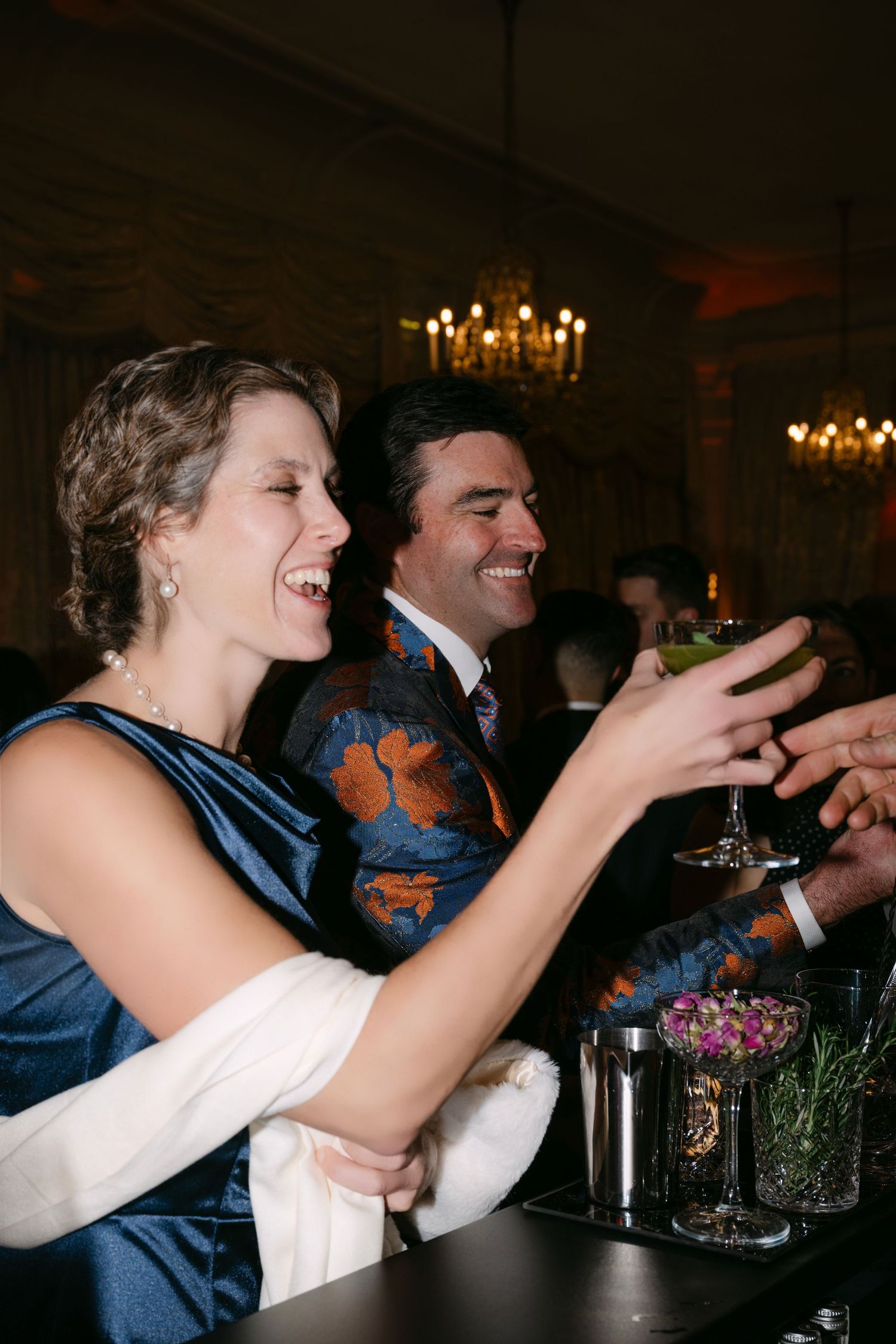 Laughing couple in elegant attire toasting cocktails at a glamorous wedding reception bar.
