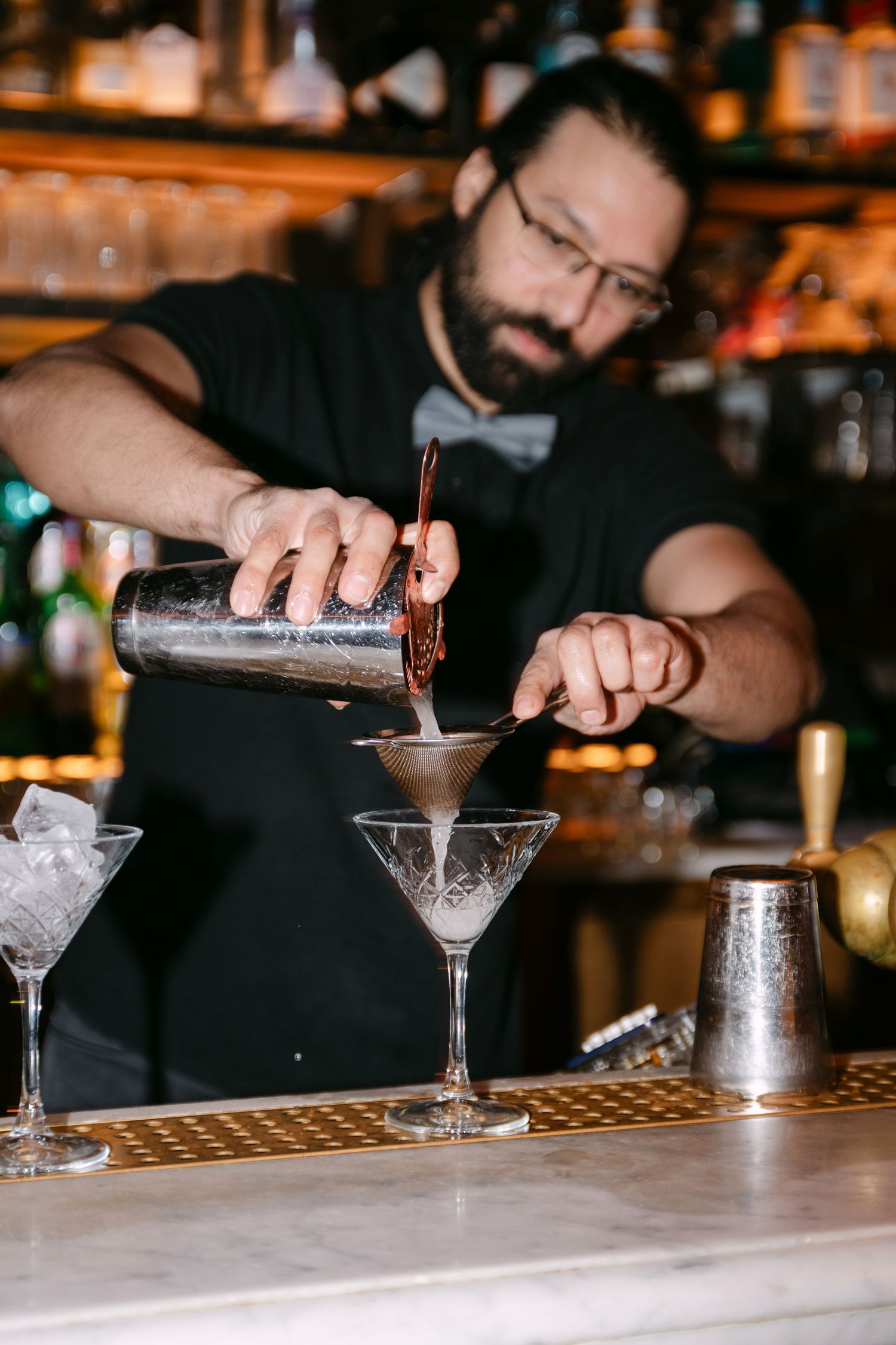 Bartender pouring cocktail through strainer into crystal martini glass at bar.