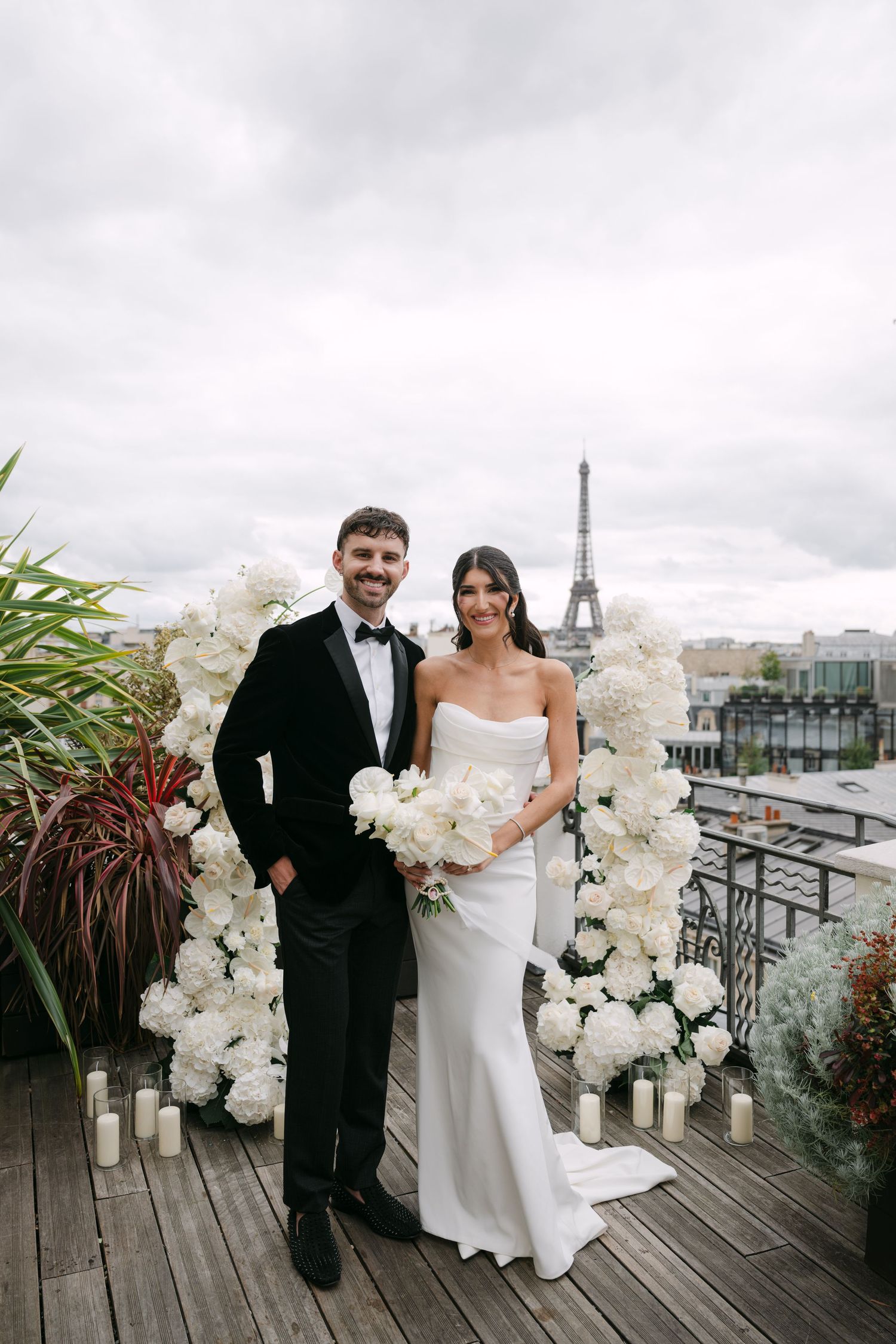 Smiling bride and groom pose before white floral arch on rooftop with Eiffel Tower in Paris.