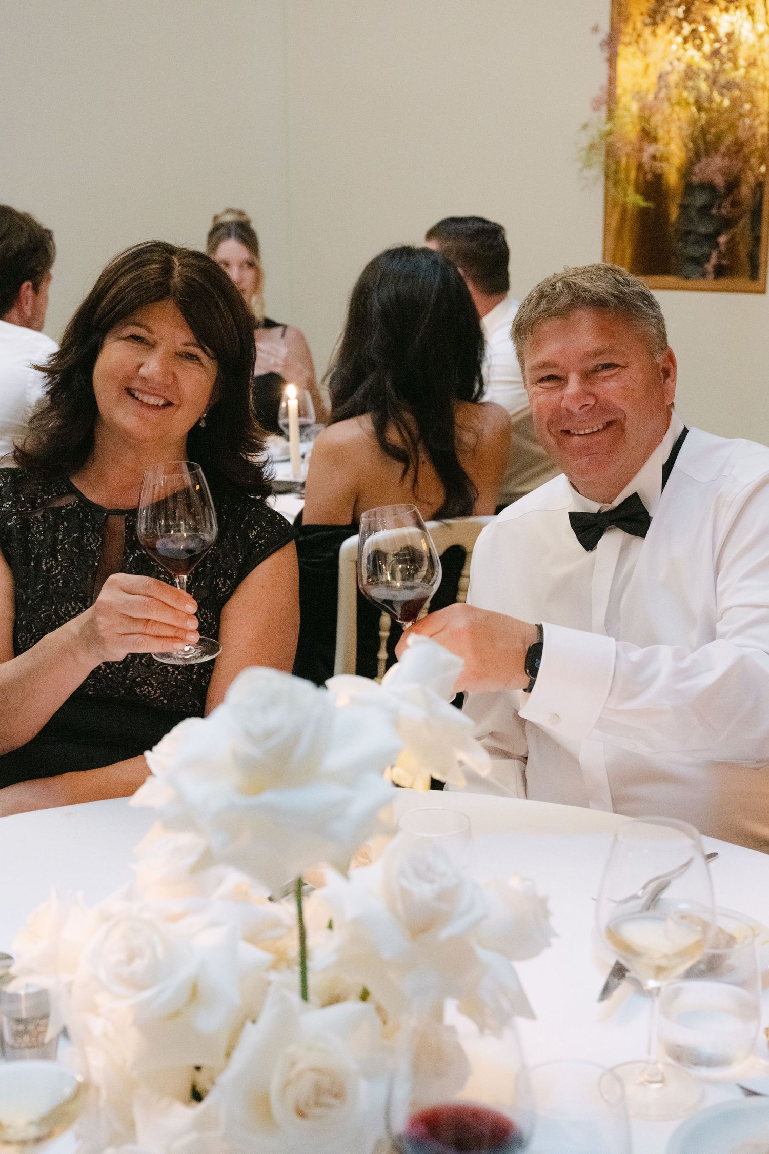 Couple toasting with wine glasses at a formal wedding reception with white roses.
