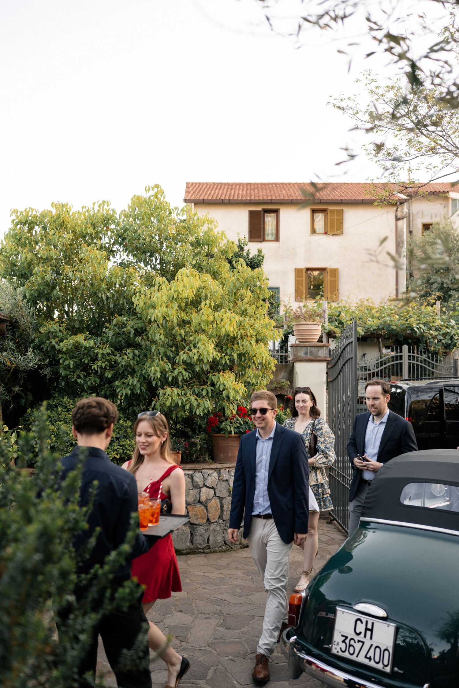 Elegantly dressed wedding guests arrive at a rustic Italian villa beside a vintage green car.