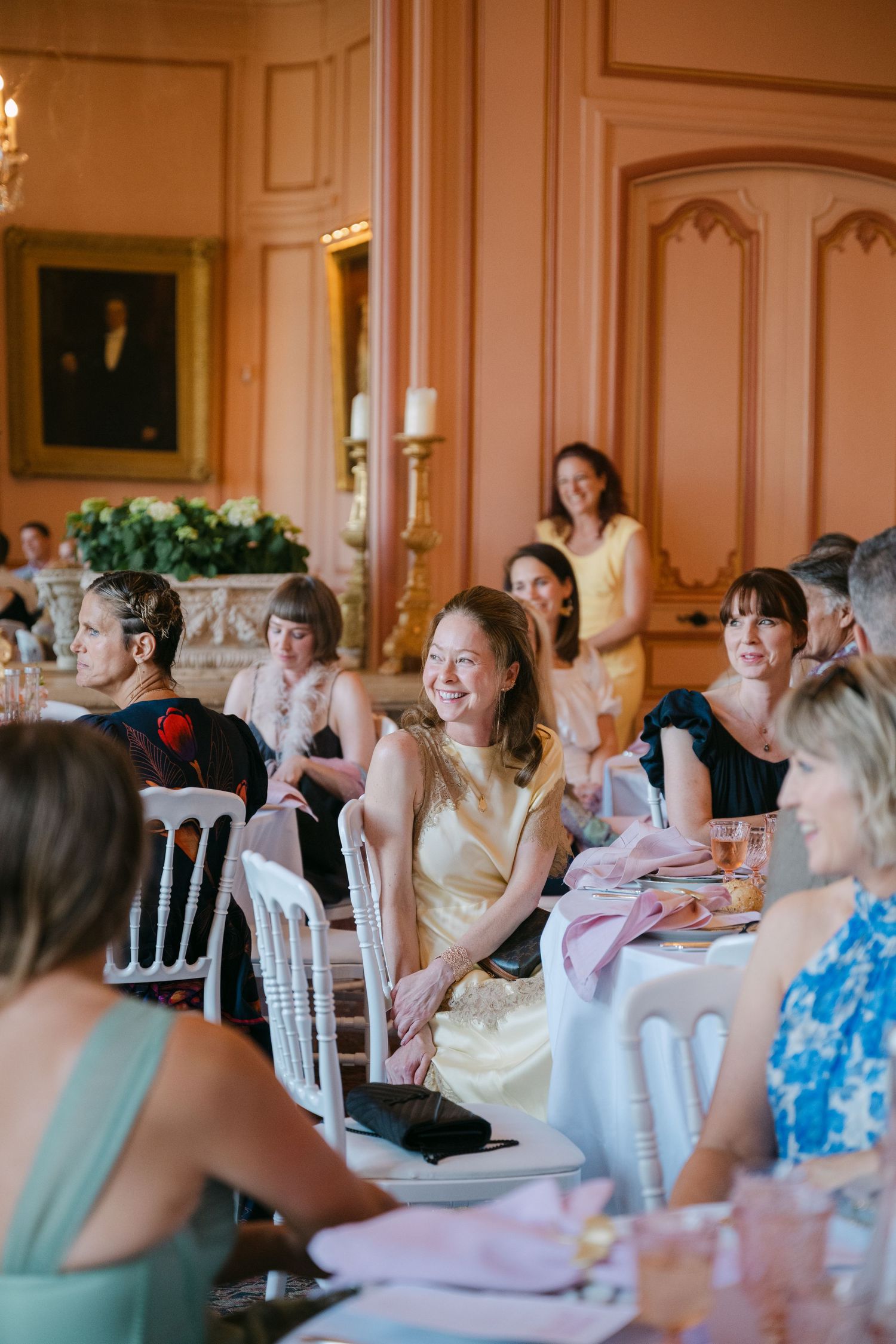 Smiling wedding guests in formal attire seated at tables in a pink-walled French chateau dining room.
