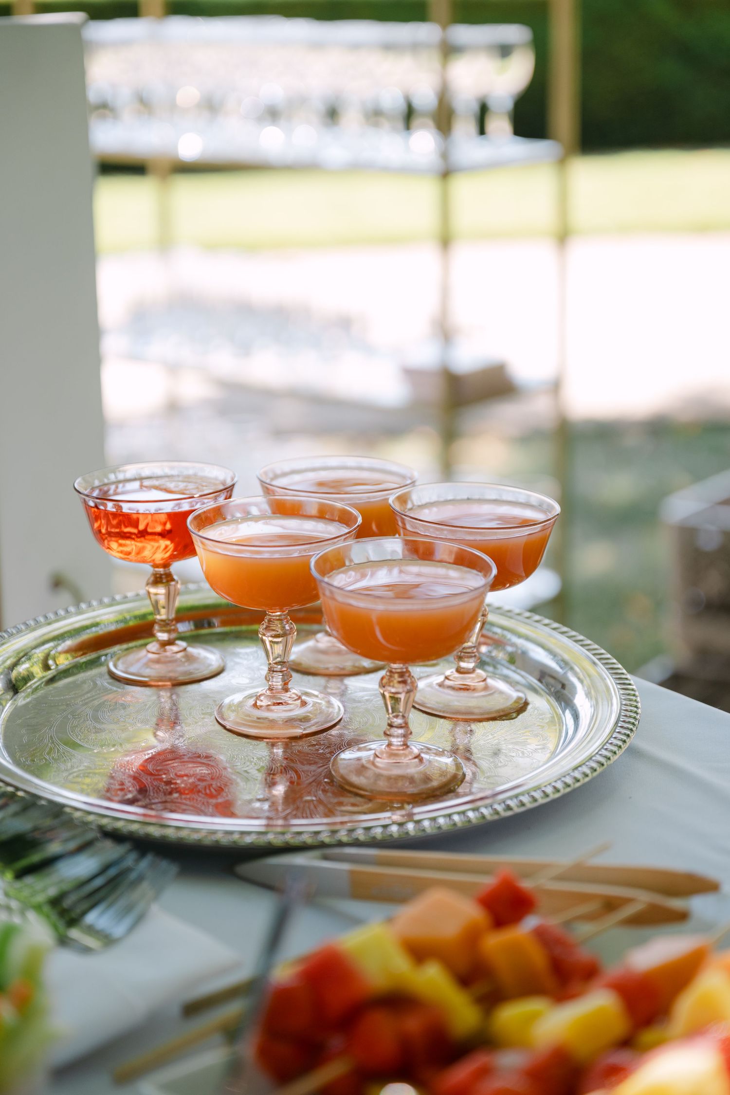 Six pink coupe glasses filled with orange cocktails arranged on a silver tray at an outdoor wedding reception.