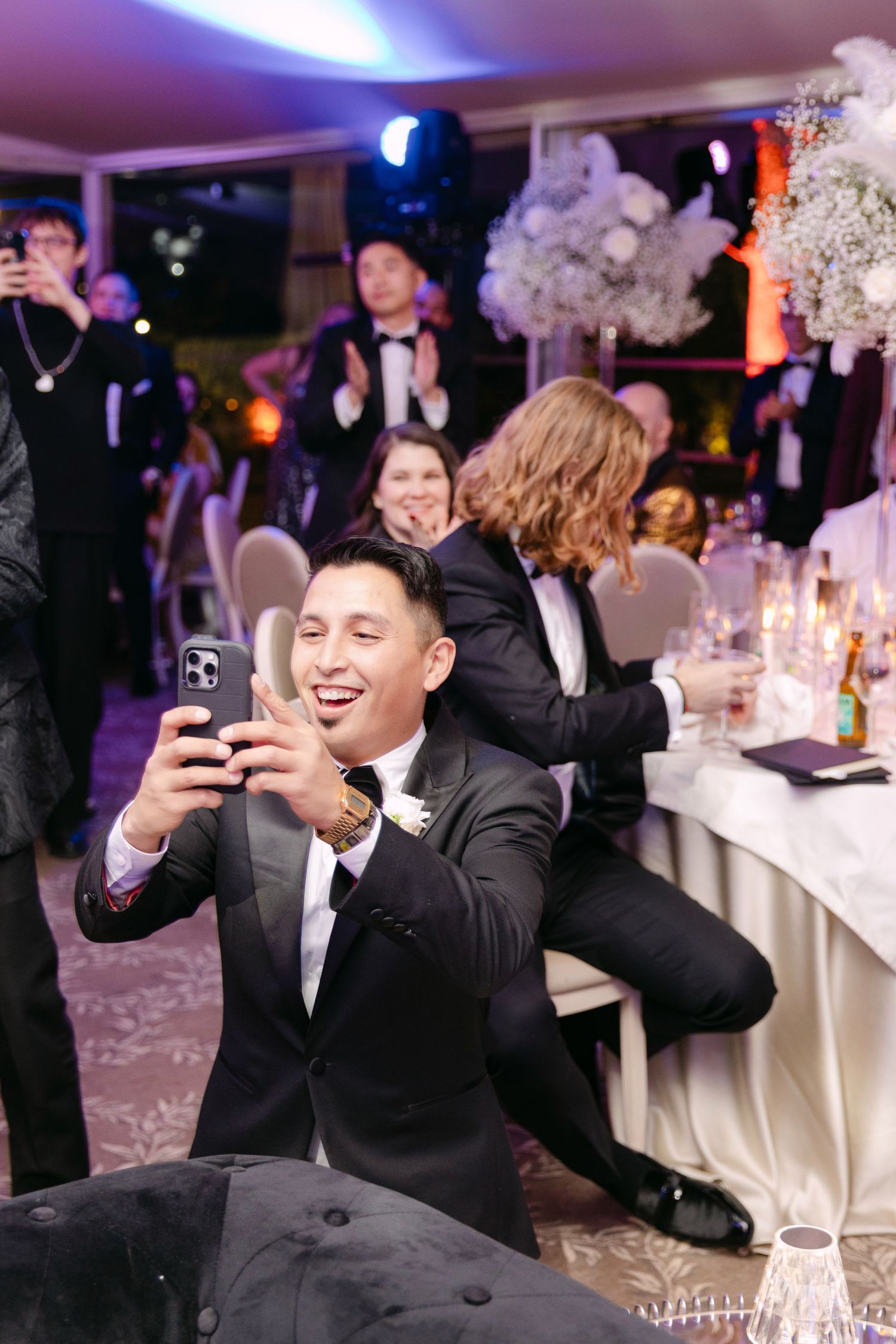 Guest in black tuxedo takes smiling selfie at lively wedding reception celebration.