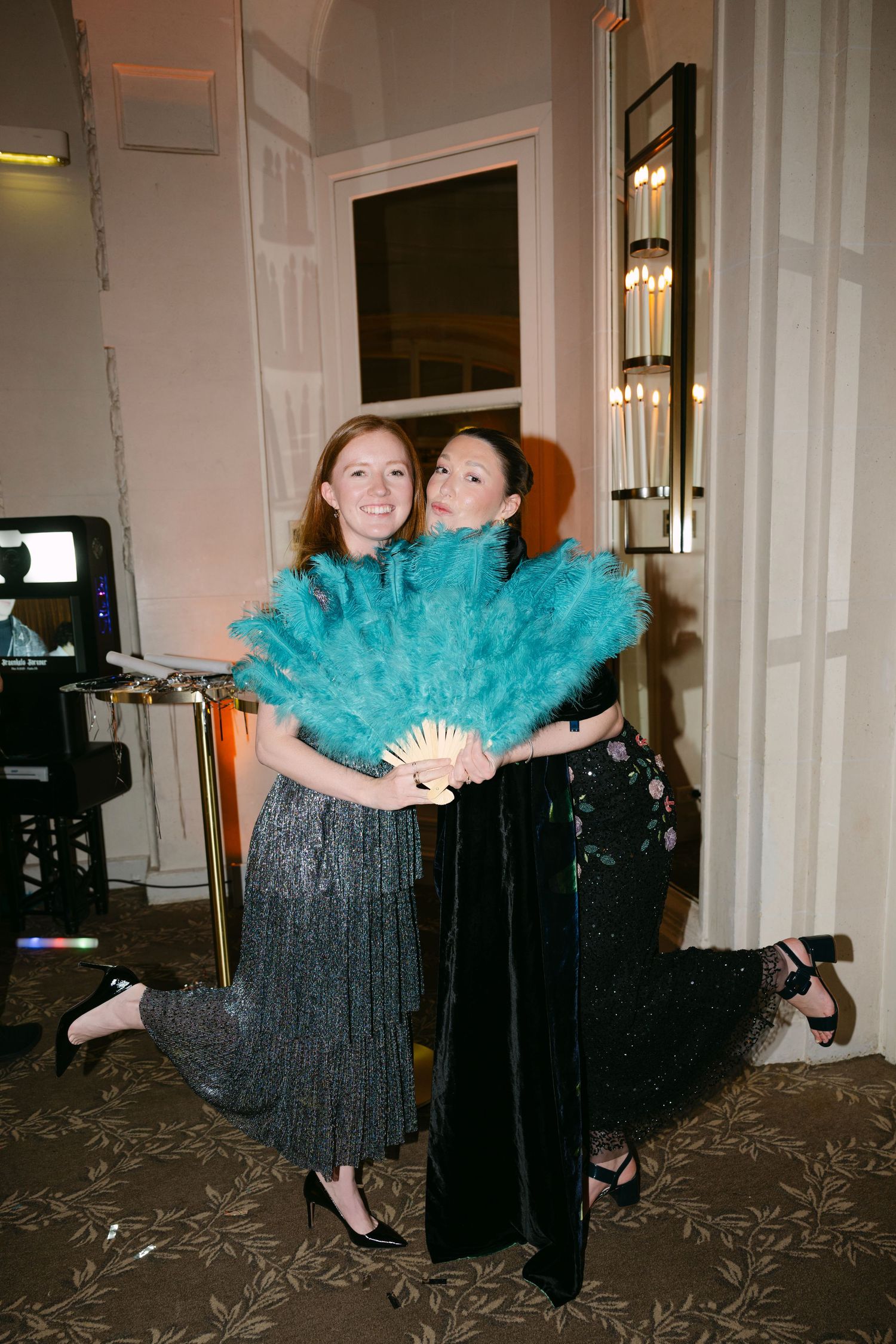 Two women posing together holding a large teal feather fan at an elegant evening event.