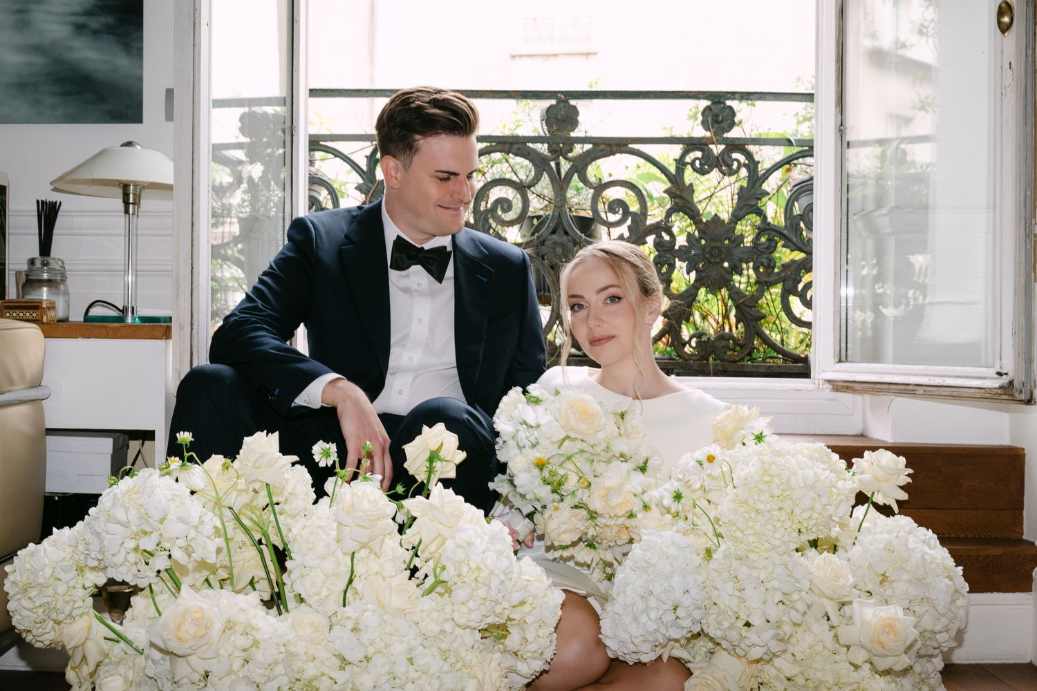 Elegant wedding couple signs documents at a table decorated with lush white floral arrangements in a bright room.