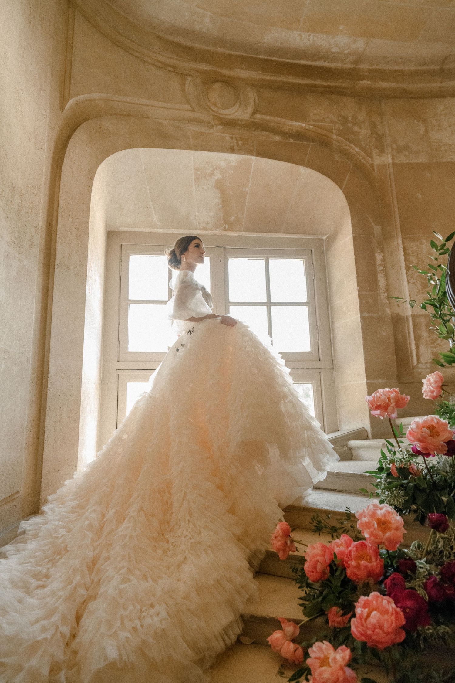 Dramatic wedding dress with flowing train photographed by window with pink roses.