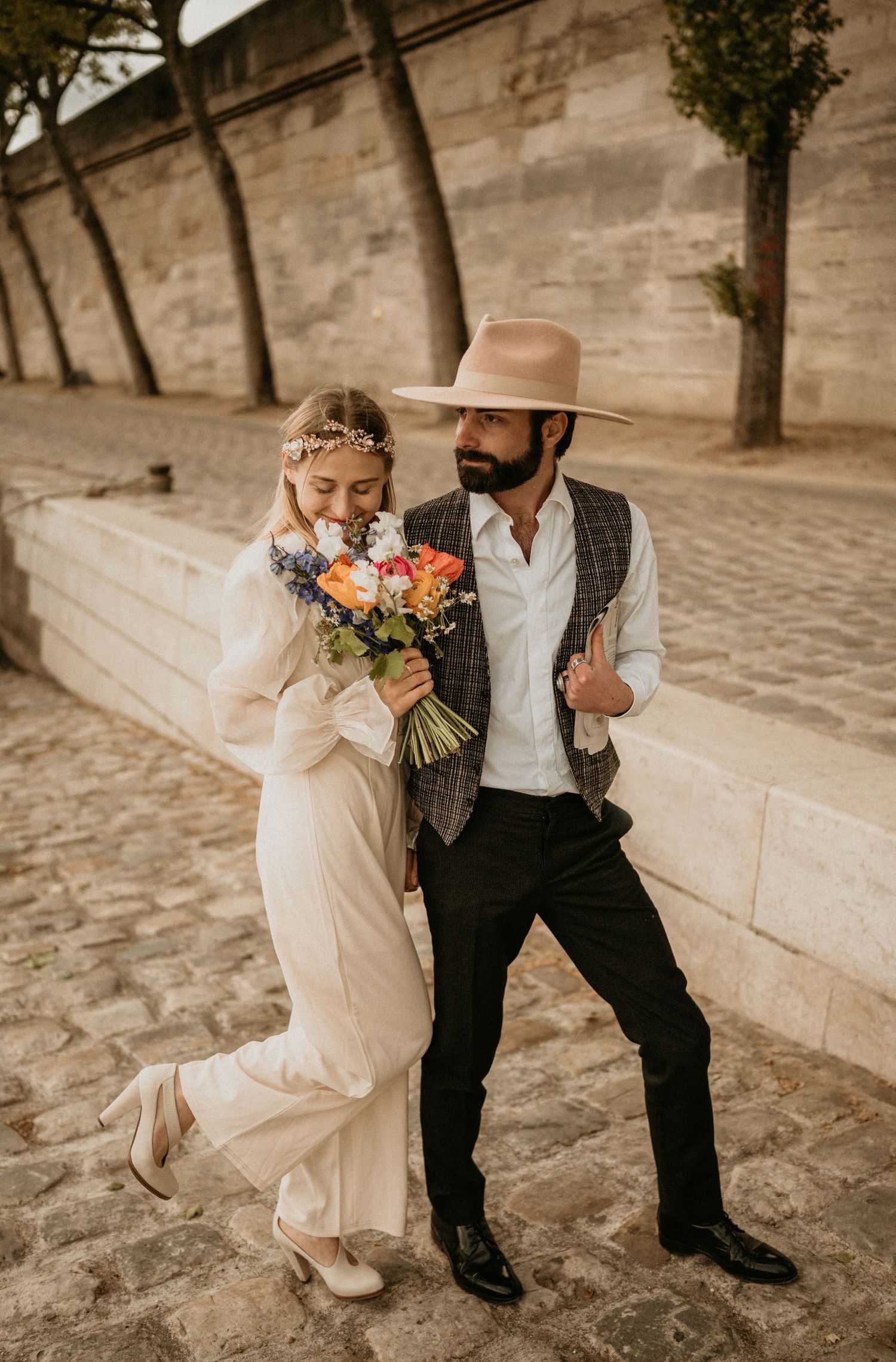 A stylish couple in wedding attire posing with a colorful bouquet against a stone wall.