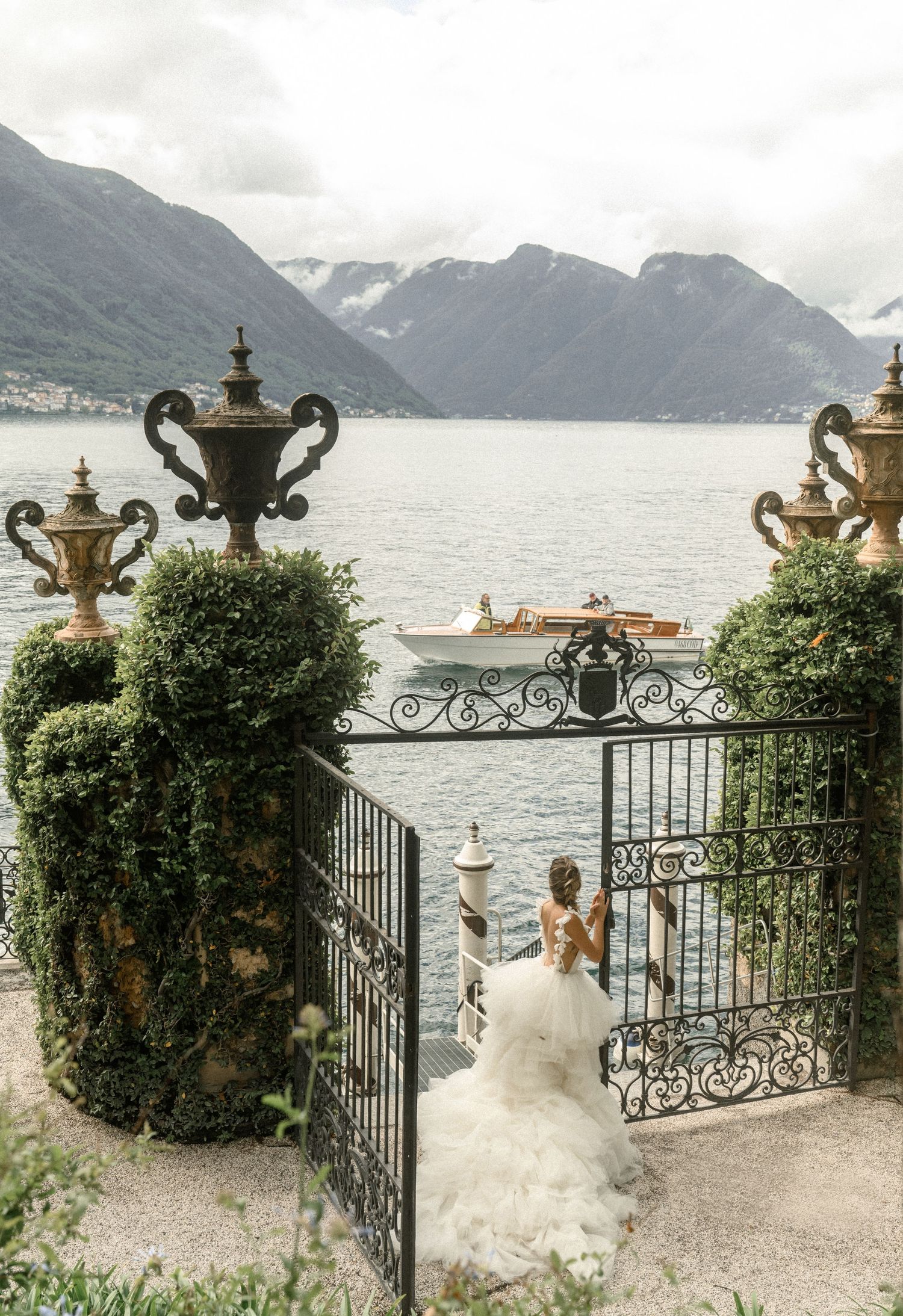 Ornate iron gates frame a scenic lake view with mountains and decorative urns in an elegant garden setting.