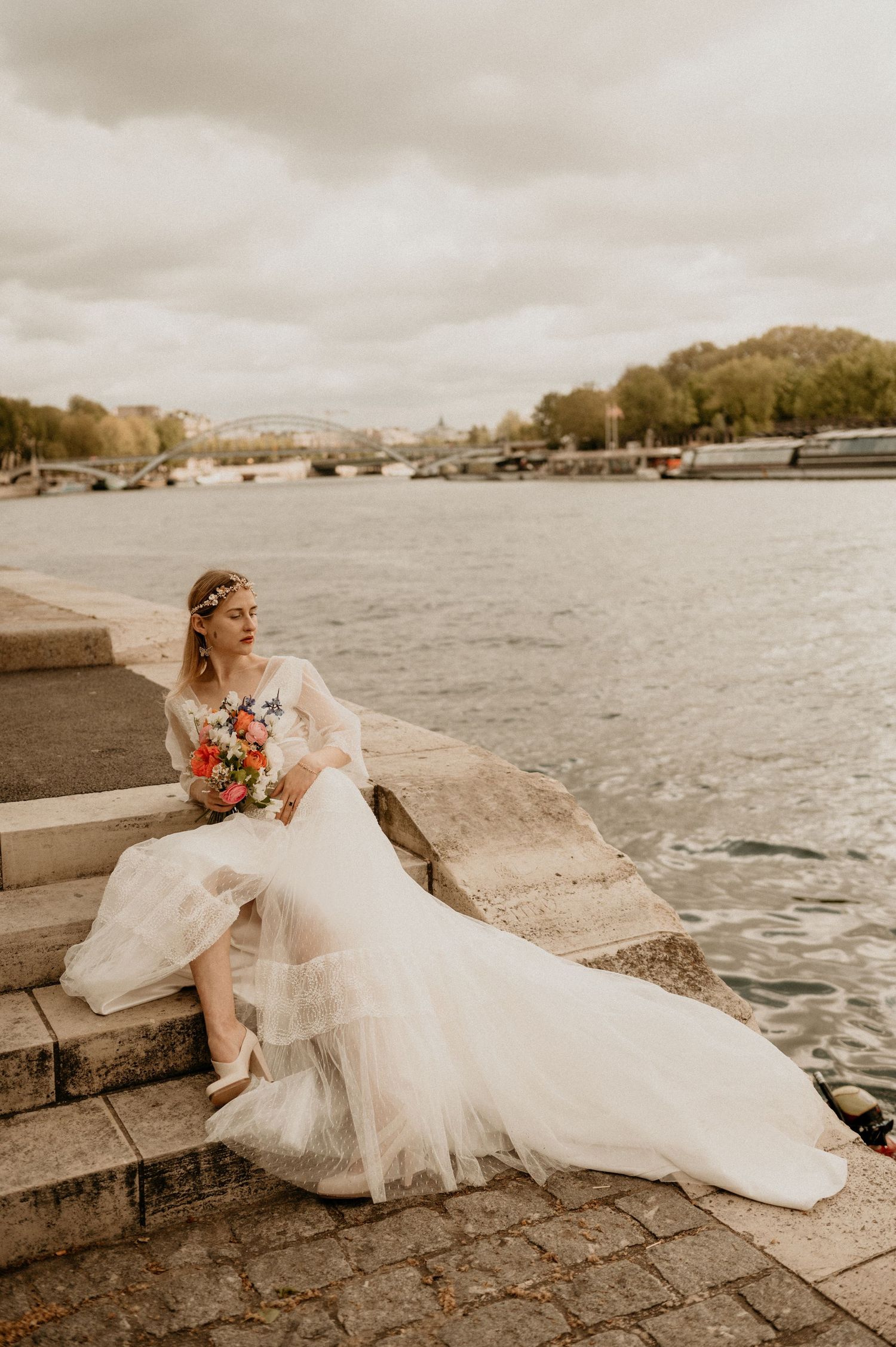A sepia-toned bridal portrait showing a bride with a bouquet seated on riverside steps.