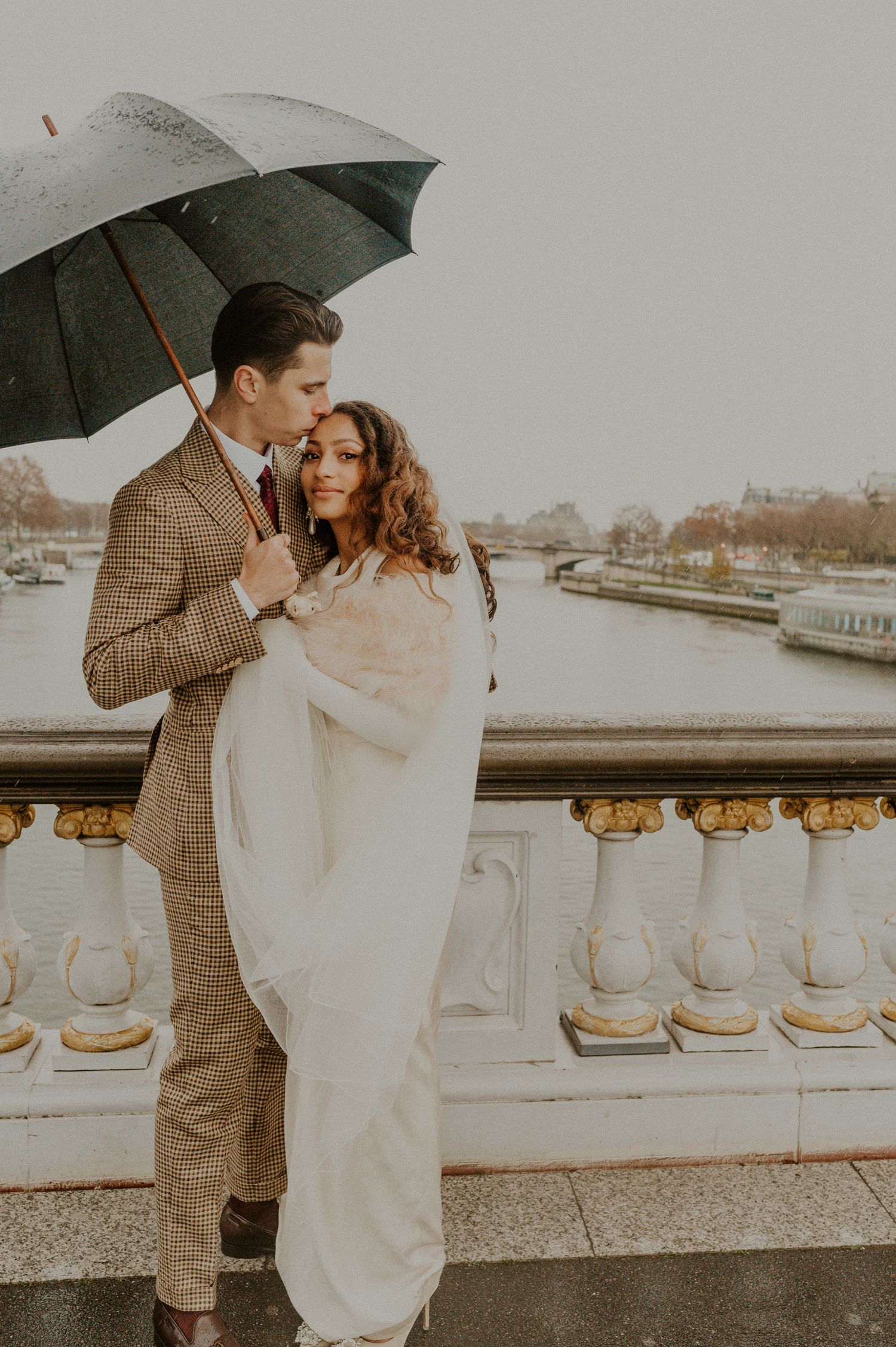 A romantic couple embraces on a stone balcony under an umbrella in Paris on a cloudy day.