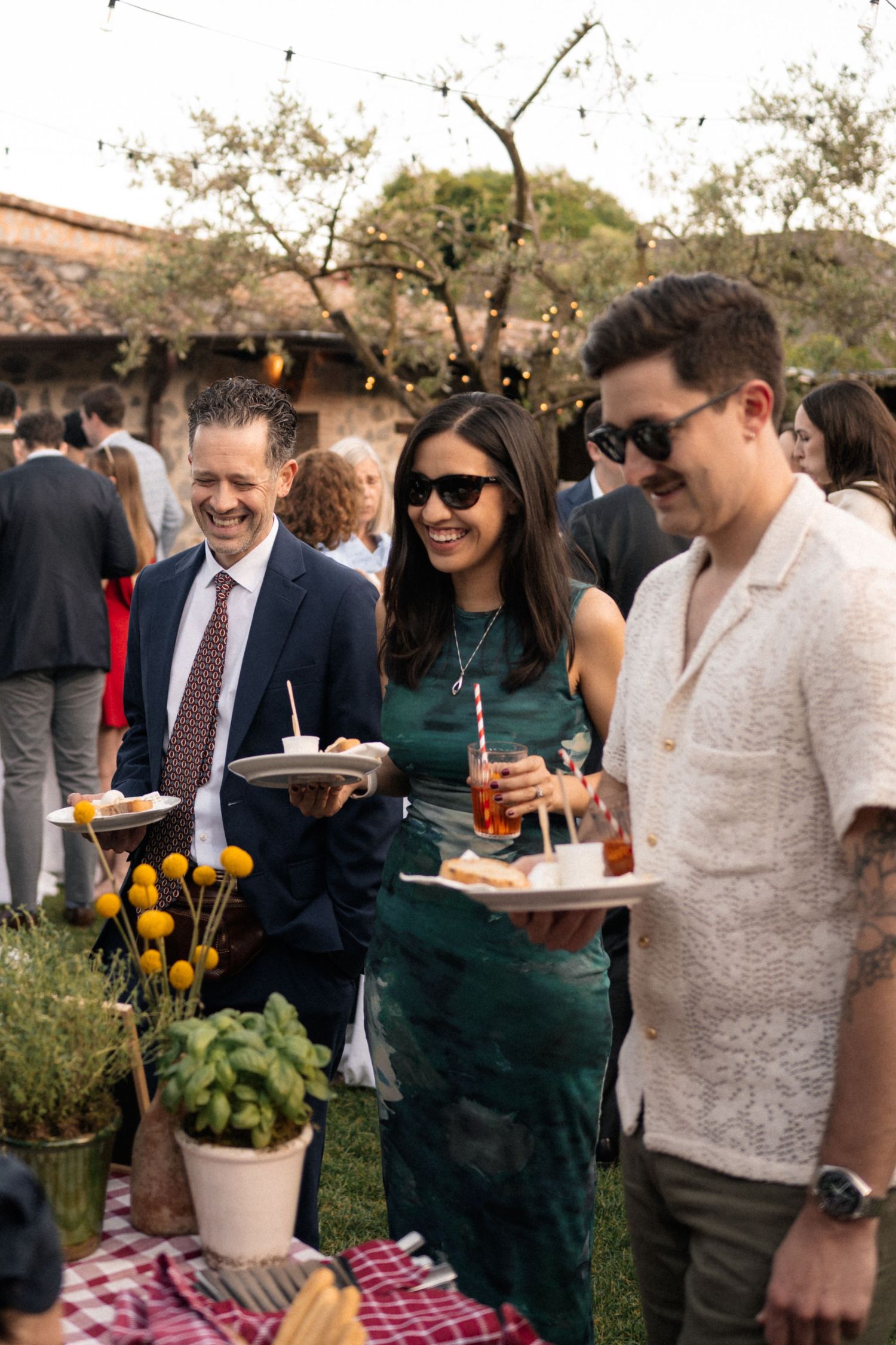 Three elegantly dressed wedding guests smiling and enjoying appetizers and drinks at an outdoor reception.