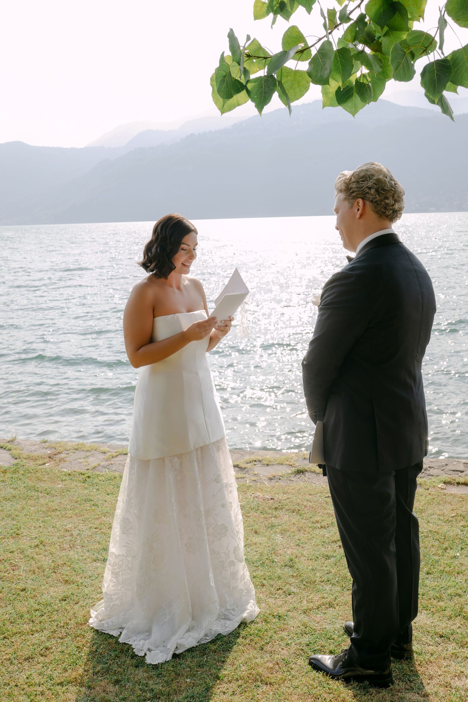 Bride in a strapless white lace gown reads her vows to her groom in a dark suit on a grassy lakeside lawn with mountains behind.