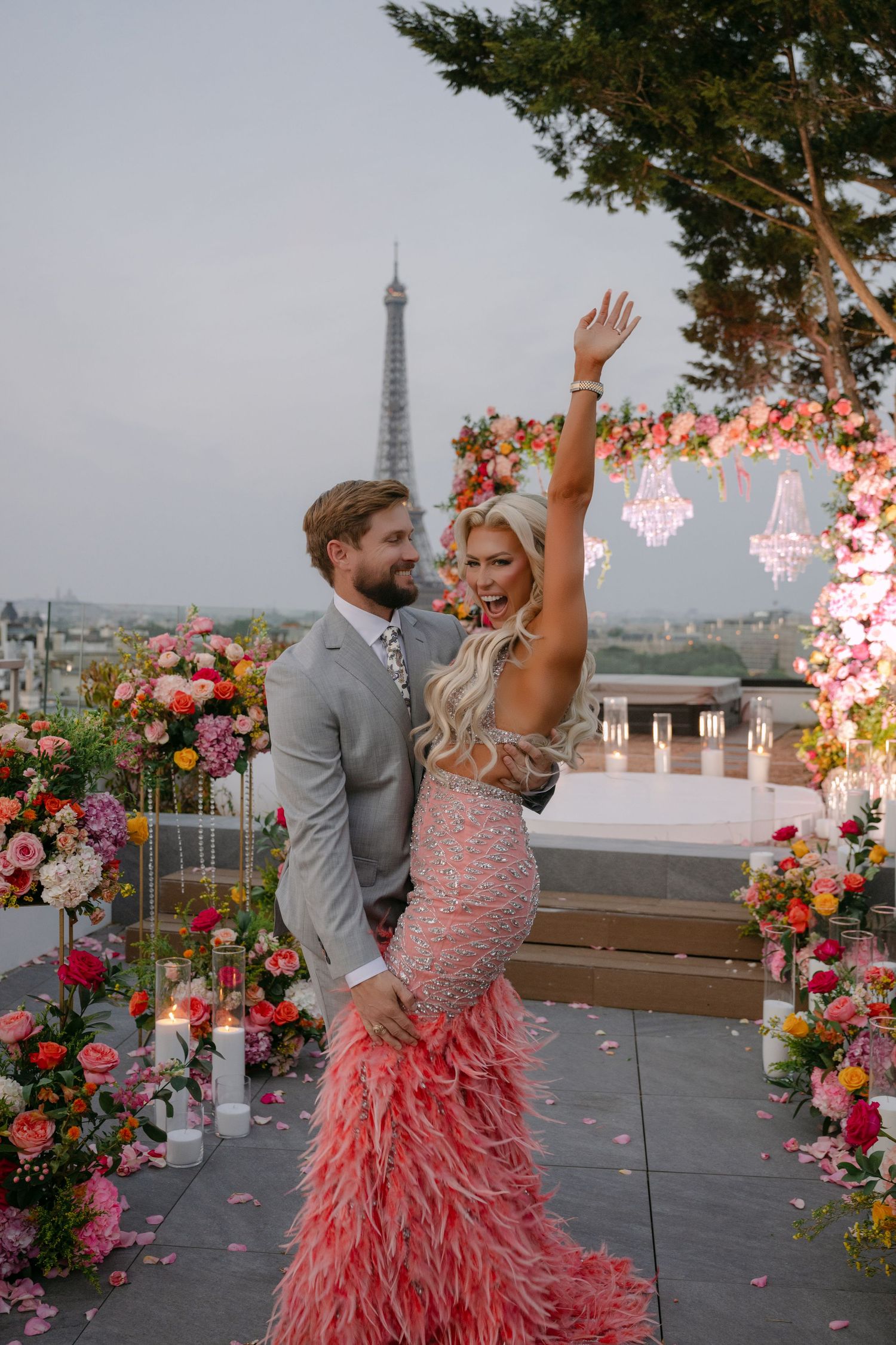 Couple celebrating in front of Eiffel Tower, woman in pink feather gown raises arm, surrounded by colorful floral arrangements.