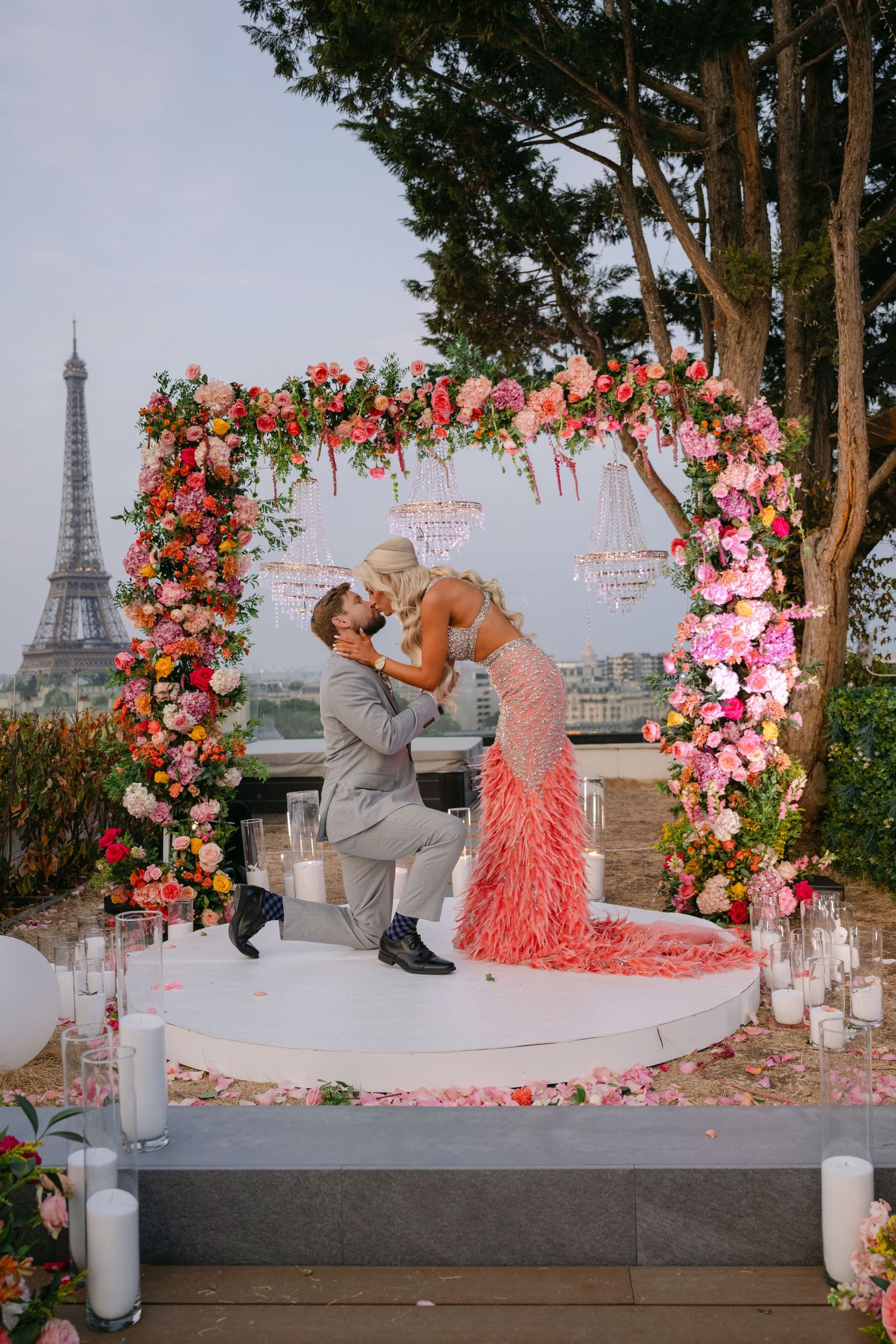 Romantic Paris proposal under floral arch with Eiffel Tower backdrop, couple kissing surrounded by pink roses and candles.