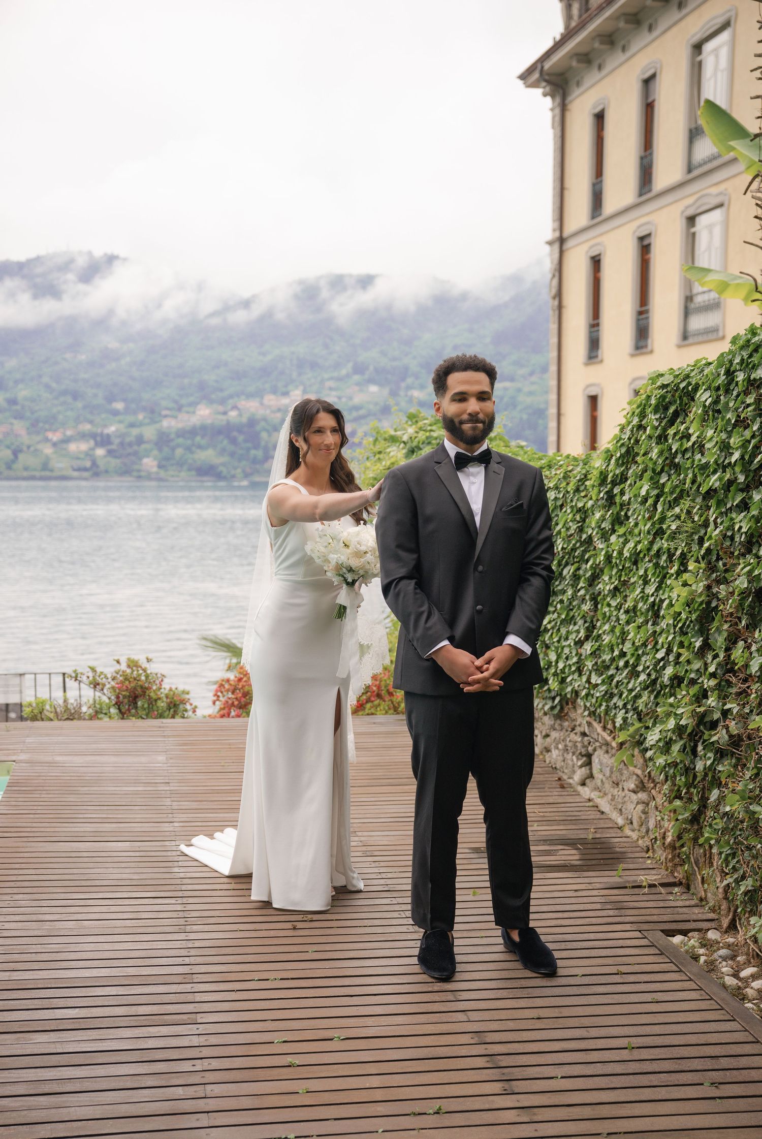 Bride in white gown with veil and groom in black tuxedo pose on wooden deck overlooking a scenic lake.