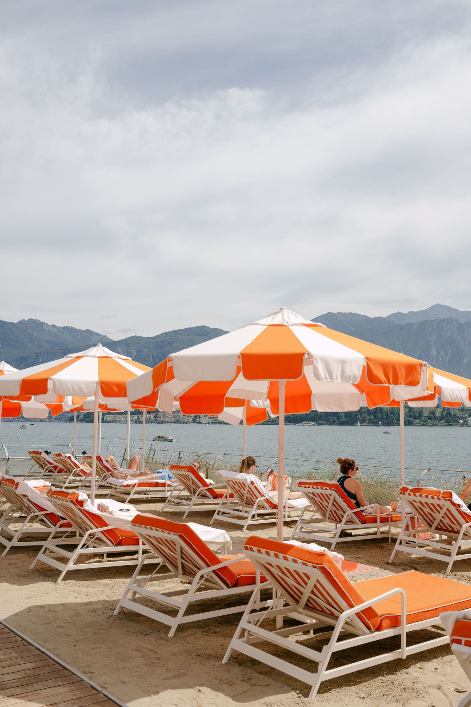 Orange and white striped beach umbrellas with matching lounge chairs by a lake, mountains in background on cloudy day.