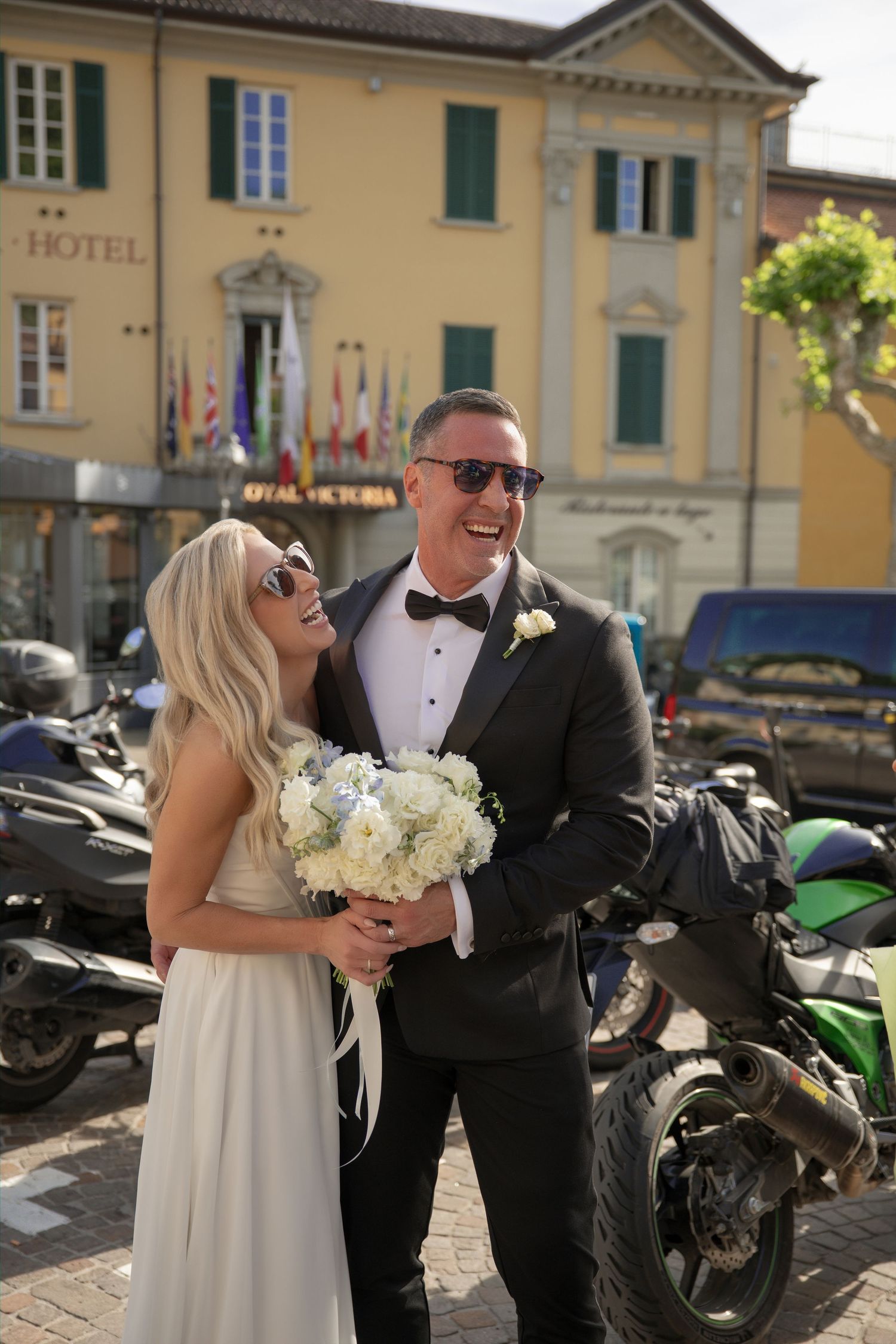 Happy bride and groom in sunglasses laughing outside a hotel in Italy, bride holding white bouquet.