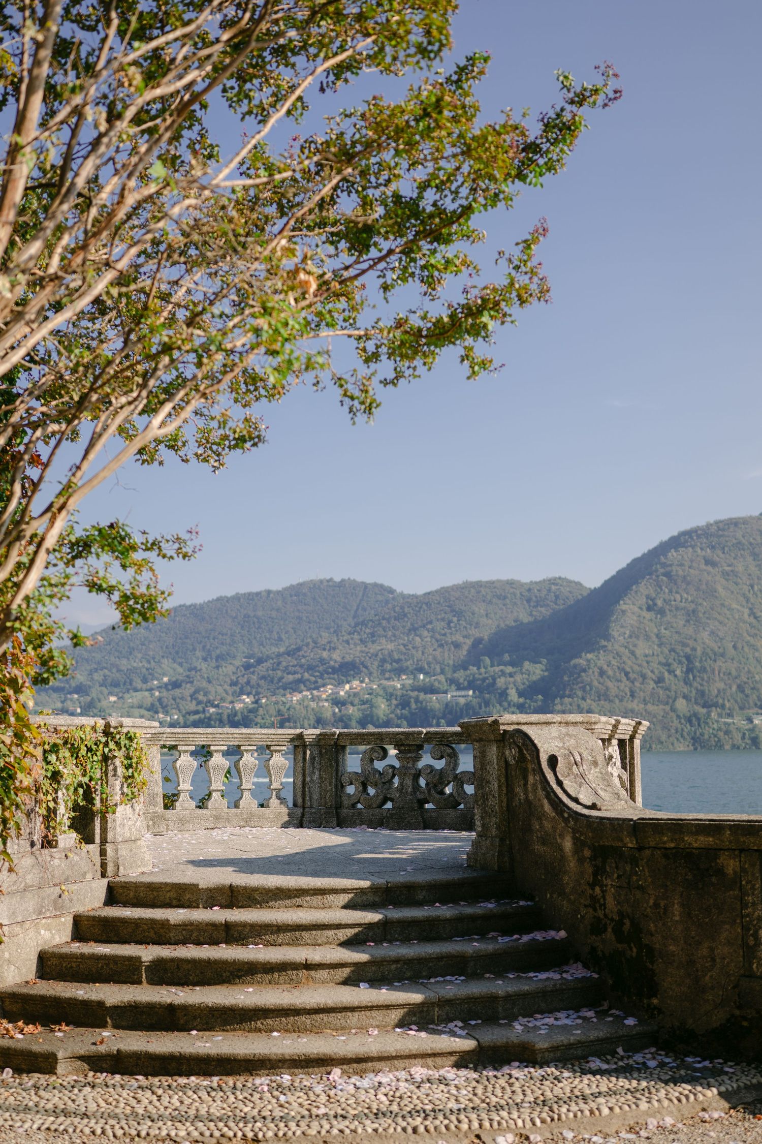 Stone terrace with ornate balustrade, curved steps, and lake and mountain views under a clear blue sky.
