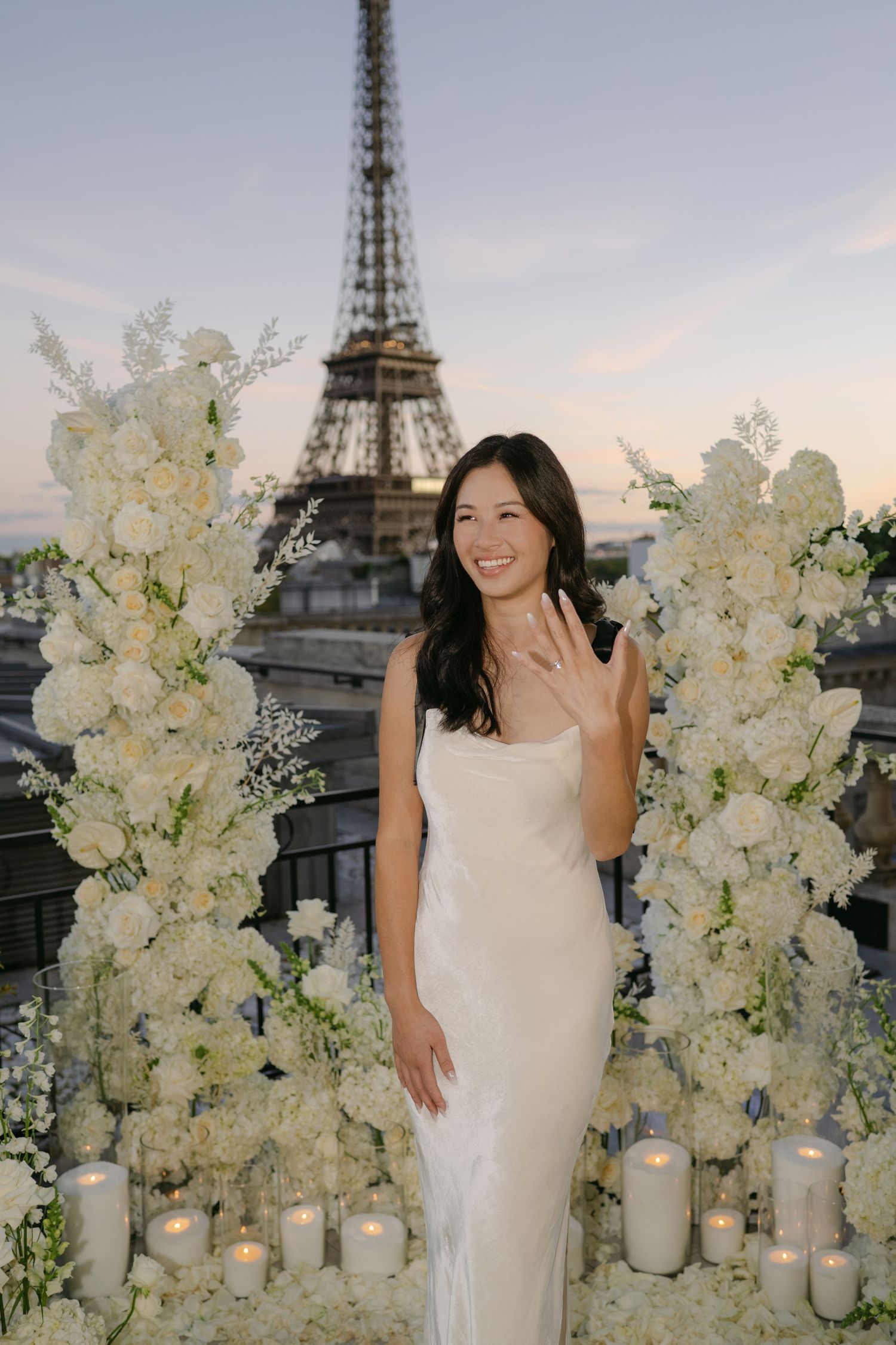 Smiling woman in a white dress shows off her engagement ring between tall white floral arches and candles with the Eiffel Tower behind.