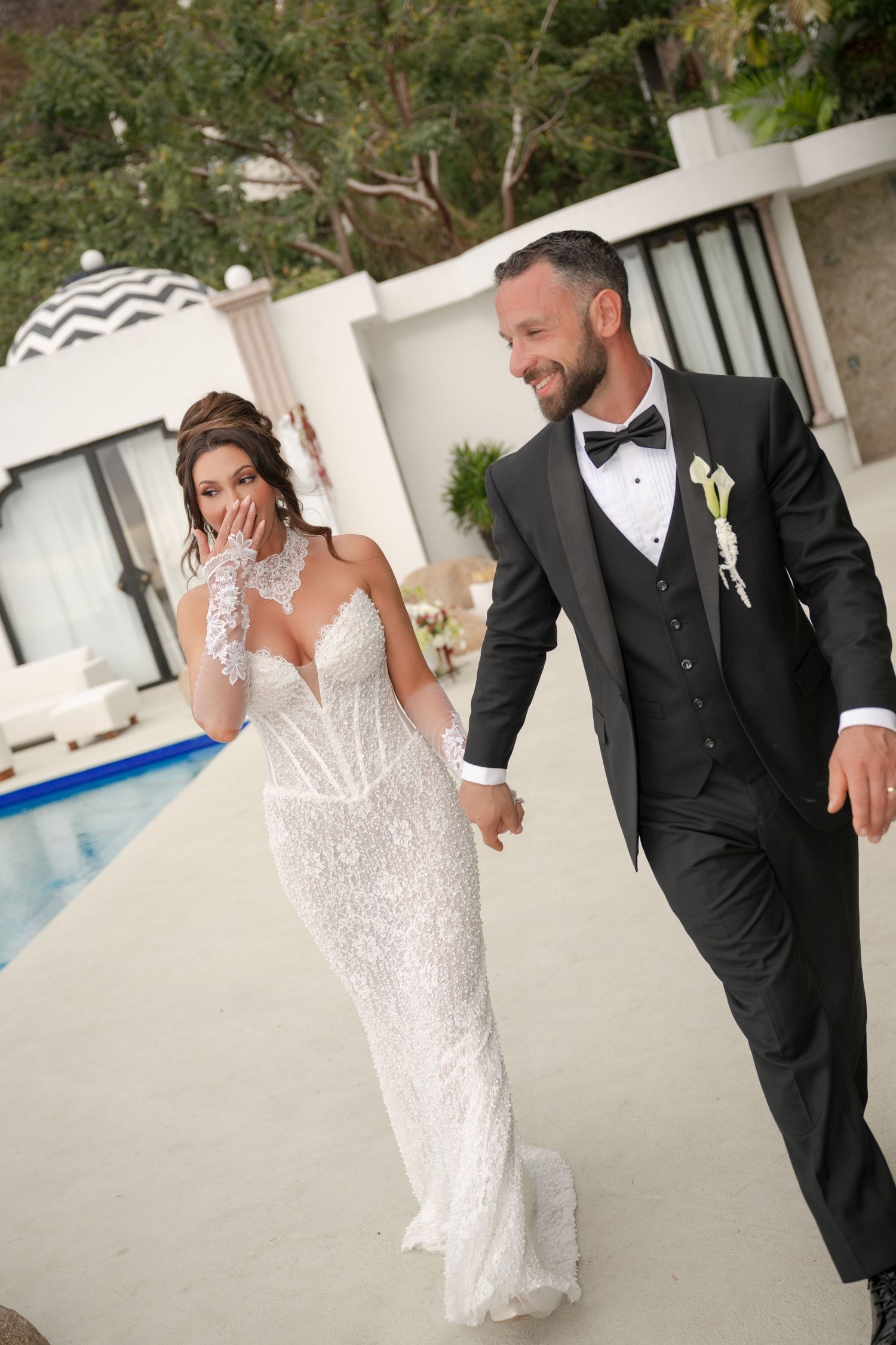 Bride in lace gown and groom in black tuxedo walking hand in hand by a pool at their outdoor wedding.