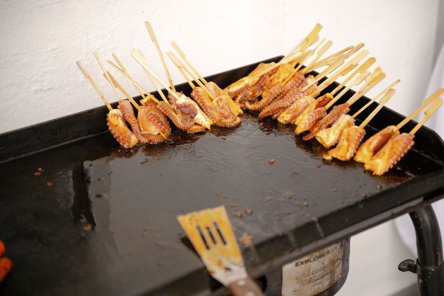 Skewered chicken satay grilling on a flat top griddle with a spatula nearby.