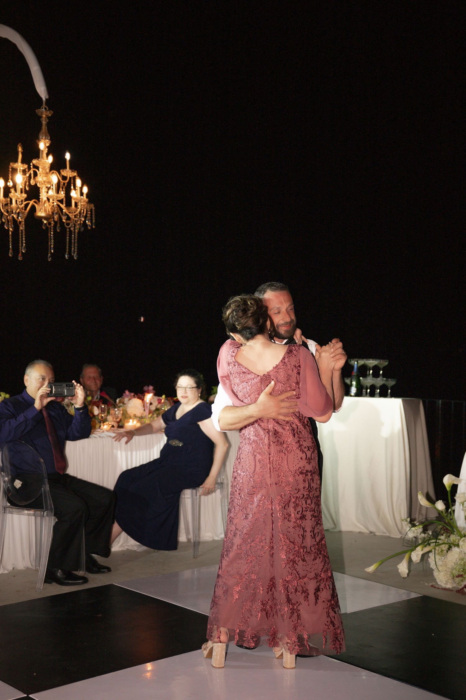 Couple sharing romantic first dance at wedding reception with chandelier and guests watching in background.