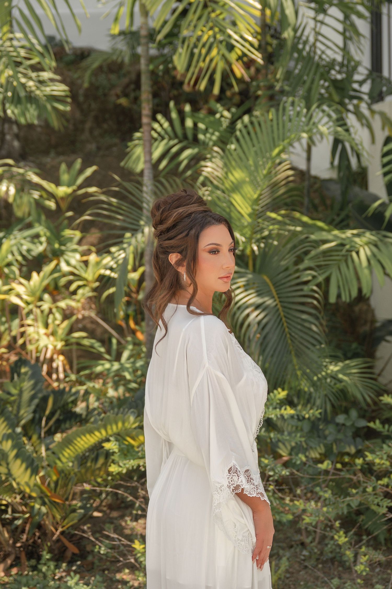 An elegant bride in a flowing white long-sleeve gown posing outdoors surrounded by lush tropical palm trees.