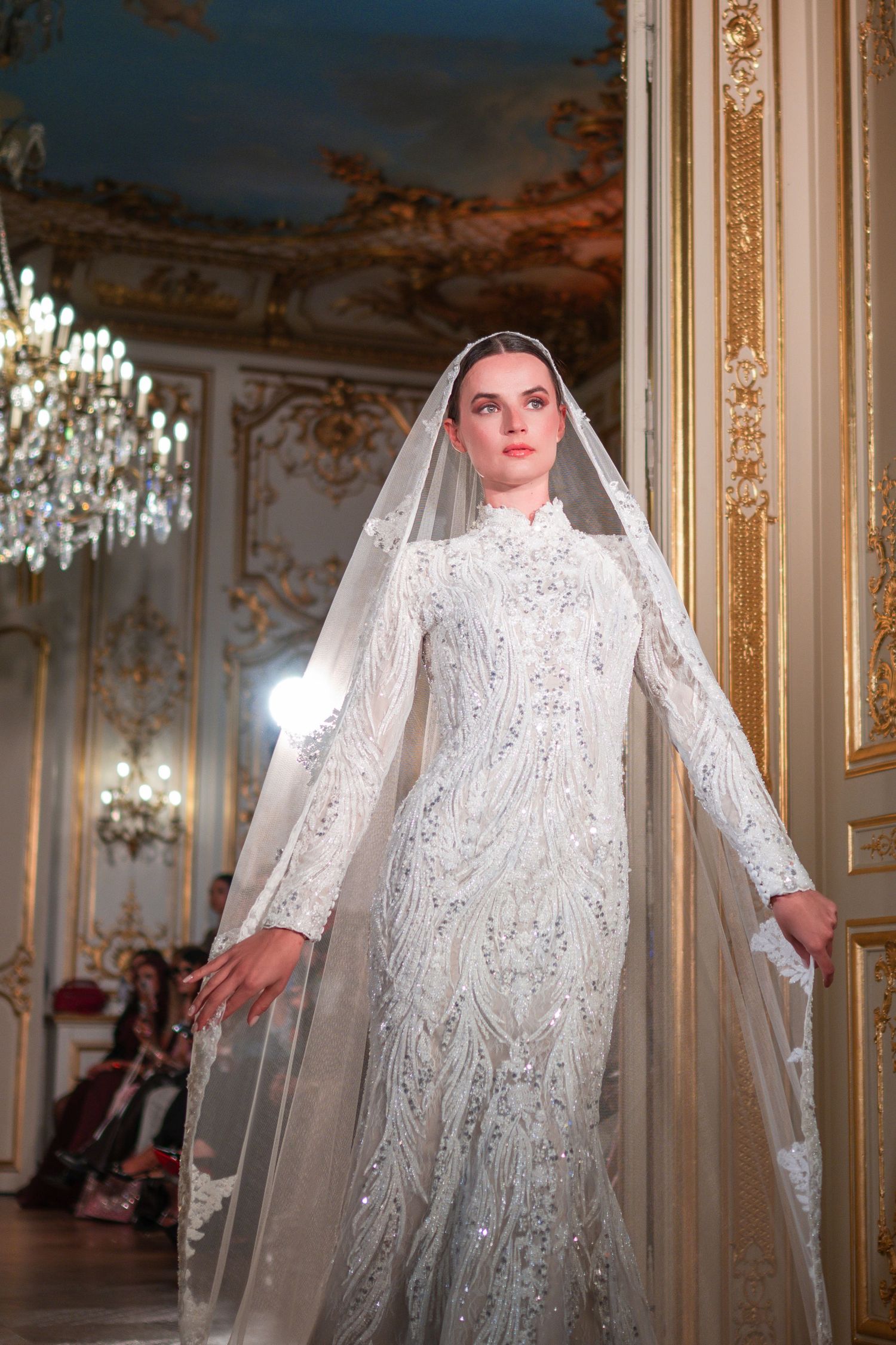 Model wearing an ornate white beaded long-sleeve wedding gown with veil in a grand gilded ballroom.