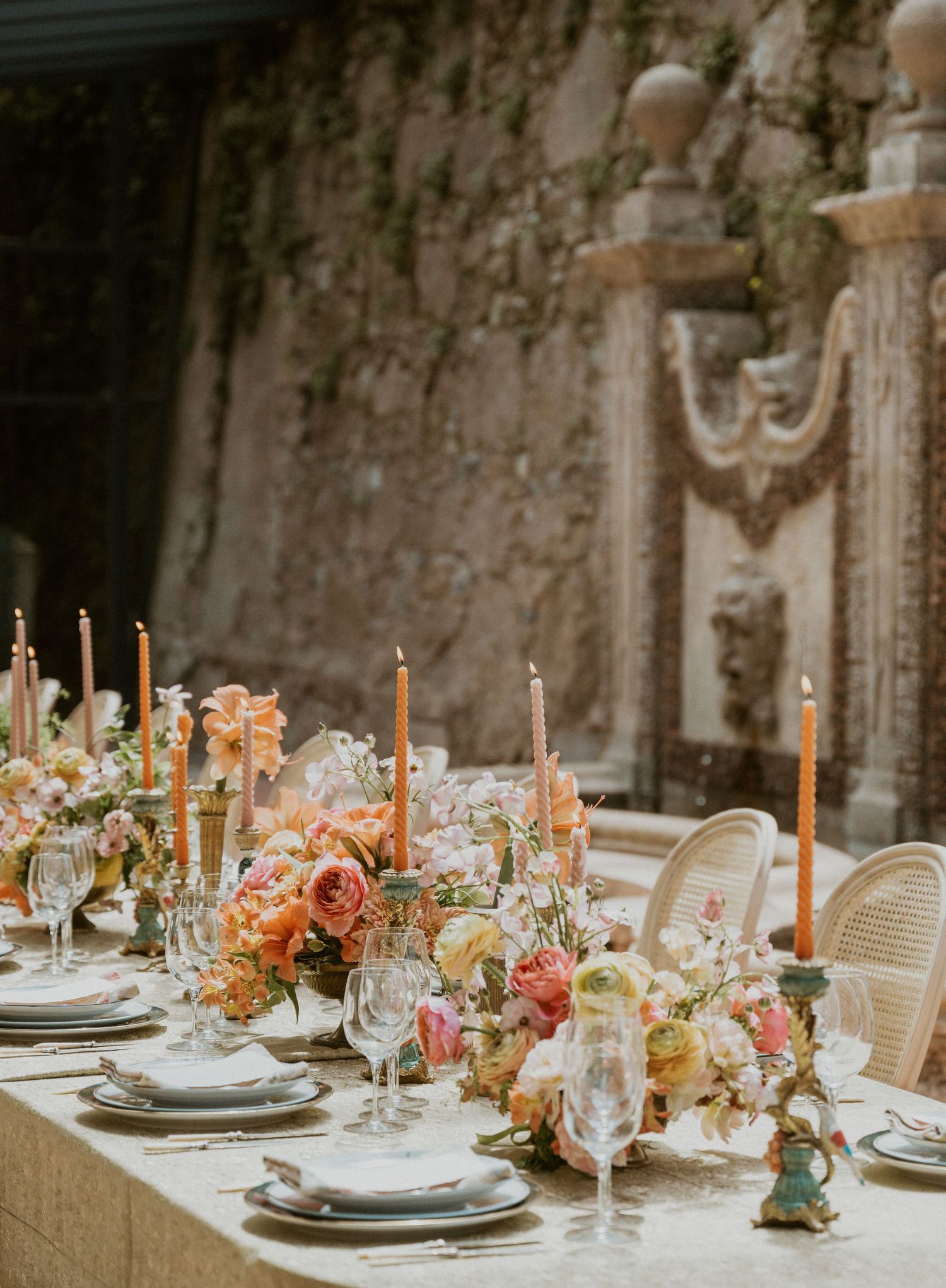 Elegant wedding tablescape with peach taper candles, lush floral centerpieces, crystal glassware, and stone wall backdrop.