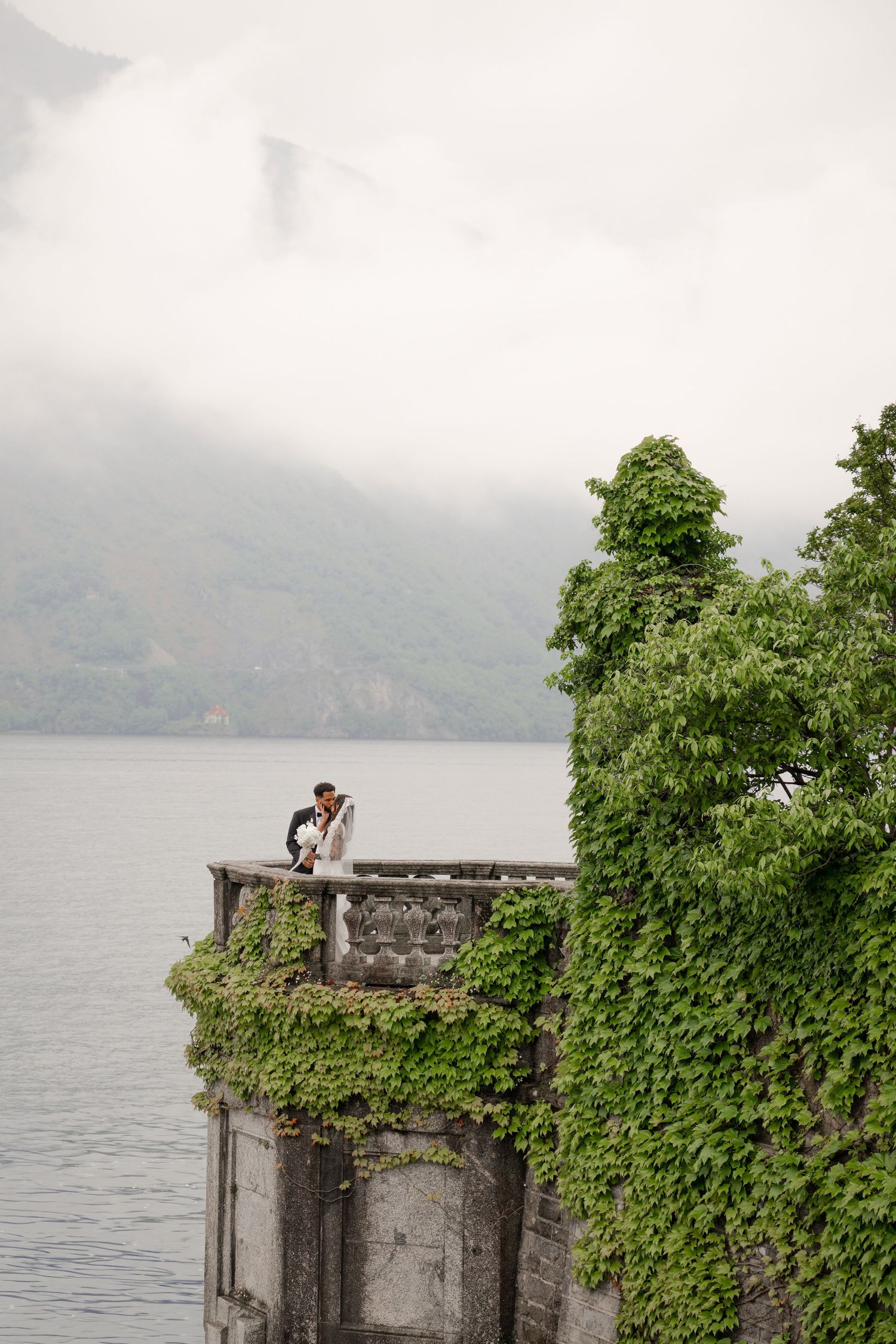 Couple embracing on ivy-covered balcony overlooking misty lake and mountains on their wedding day.