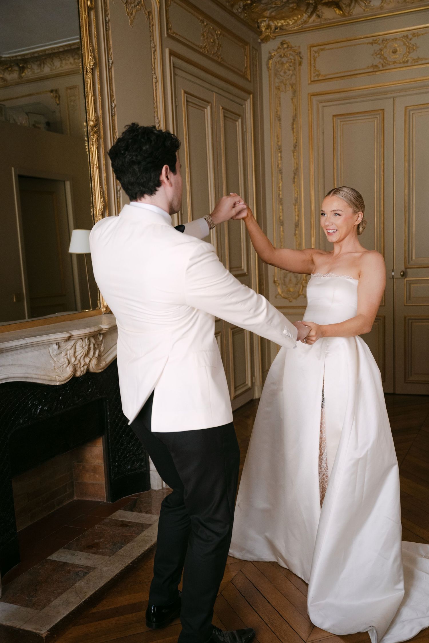 Bride and groom share a romantic dance in an elegant gold-adorned room, she in a strapless white gown, he in a white tuxedo.